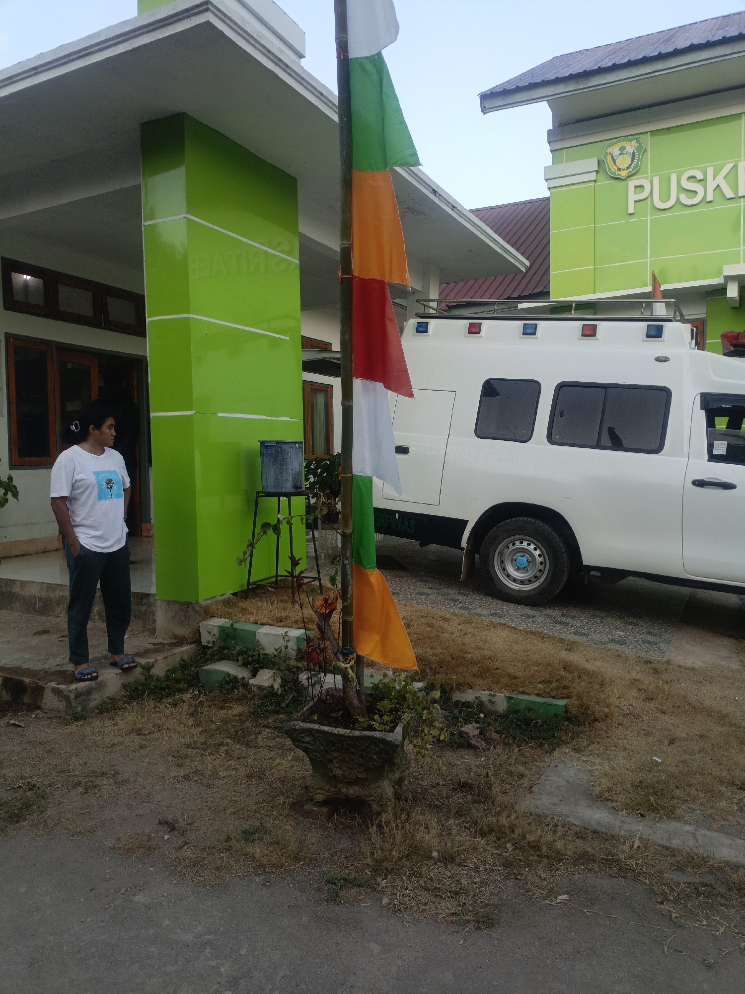 In Solor Barat, Indonesia, a white ambulance is parked in front of a bright green building with the word "PUSKESMAS" written on it. A person in a white t-shirt and dark pants stands near the entrance of the building, which features large green pillars. A flagpole with orange, white, and green flags is positioned in front of the ambulance and building. The ground is covered in dry grass and gravel. The sky is overcast, indicating it is likely early morning or late afternoon.