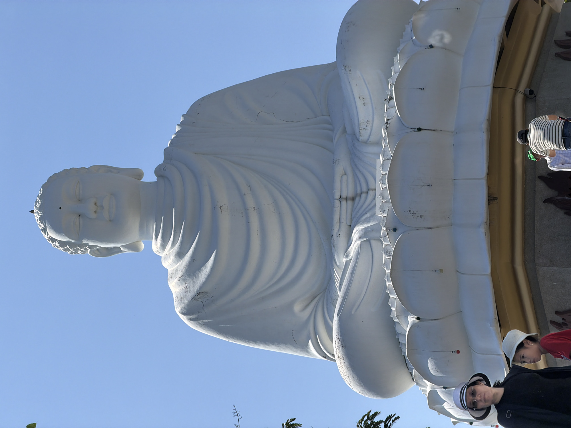 The image shows a large, reclining white Buddha statue against a clear blue sky. The statue is positioned on its left side. The statue is resting on a lotus flower base. Some people are looking at the Buddha statue at the bottom right. The statue is located in the city of Tay Nha Trang Ward, Vietnam.