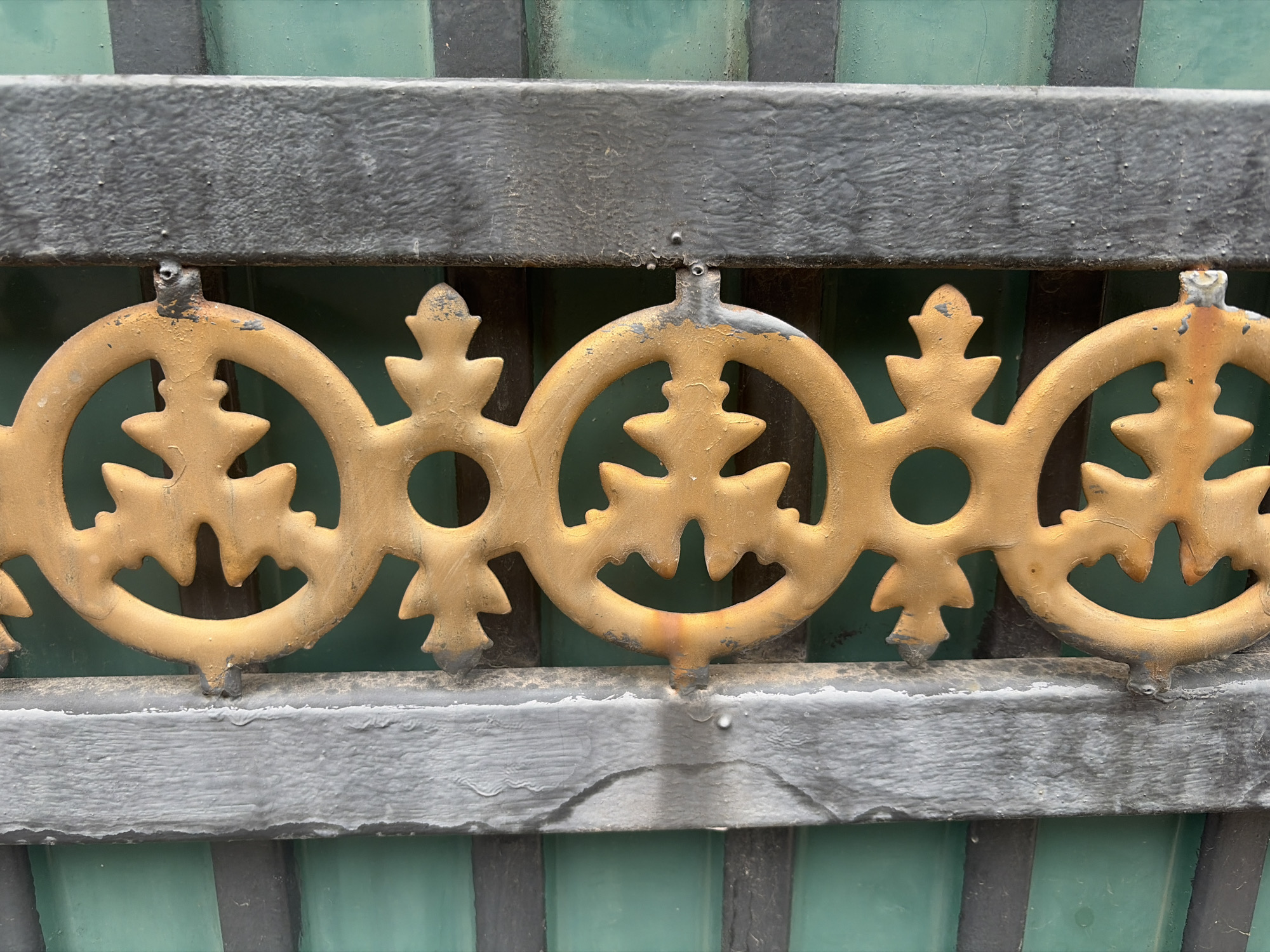 The image shows a close-up, eye-level, horizontal shot of a section of a wrought iron gate in Almaty, Kazakhstan. The gate features decorative, golden, circular elements with stylized leaf-like protrusions. These golden elements are attached to a dark grey, weathered metal bar that runs horizontally across the center of the frame. Below this bar, another dark grey, weathered metal bar is visible, also running horizontally. Behind the gate, there are vertical elements painted in alternating shades of dark green and black. The overall impression is of an old, possibly decorative, but somewhat deteriorated metal fence or gate. There are no people or animals visible in the image. The lighting suggests it is daytime. The specific location in Almaty is not identifiable from the image itself.