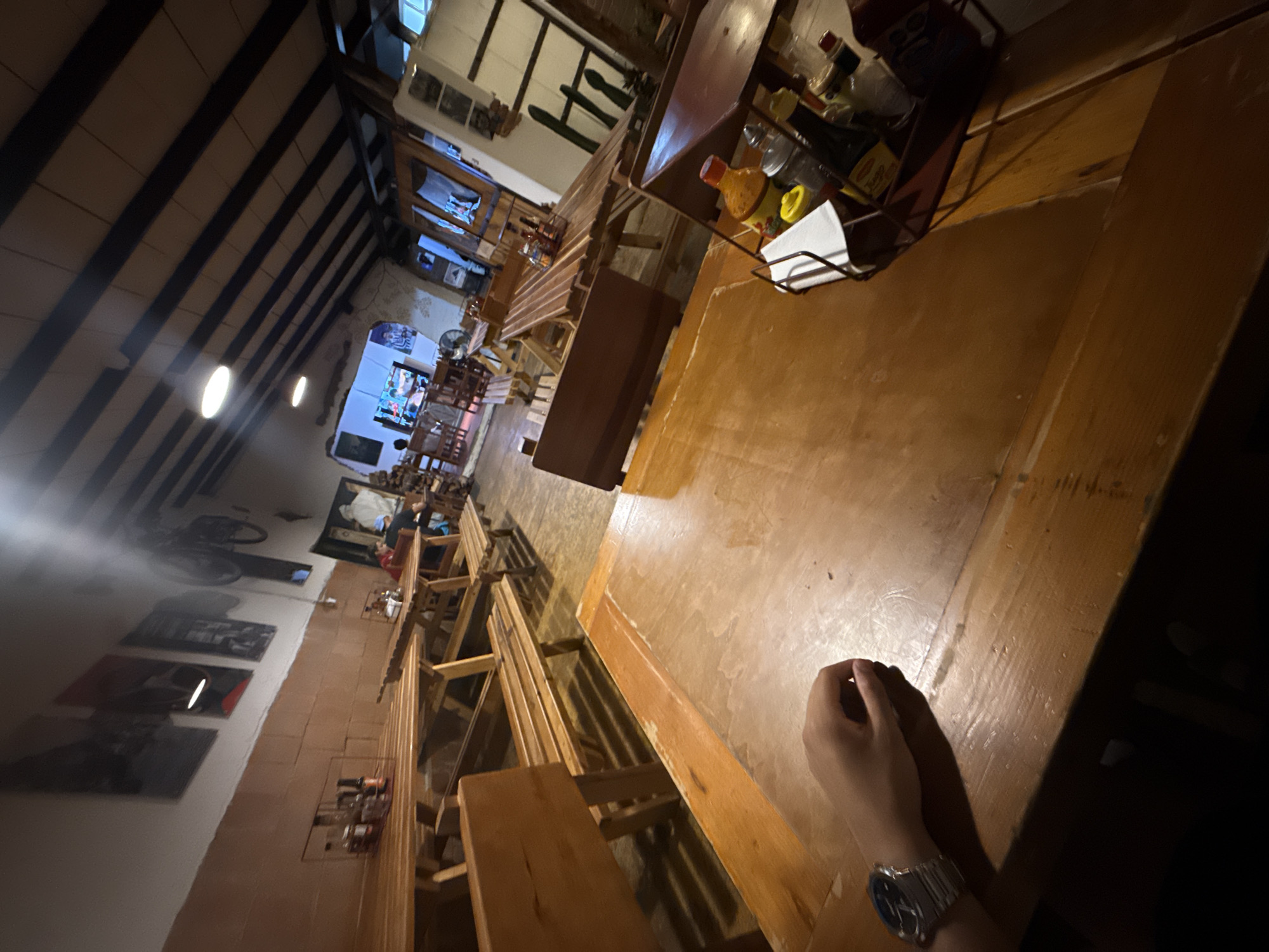 A restaurant interior in Chipilo de Francisco Javier Mina (Chipilo), Mexico. The scene captures the perspective of a person seated at a long wooden table. There are other wooden tables and benches lining the sides, stretching back into the distance. Condiment containers sit on the table. The ceiling features exposed beams. A person's hand and wrist are visible in the foreground wearing a watch.
