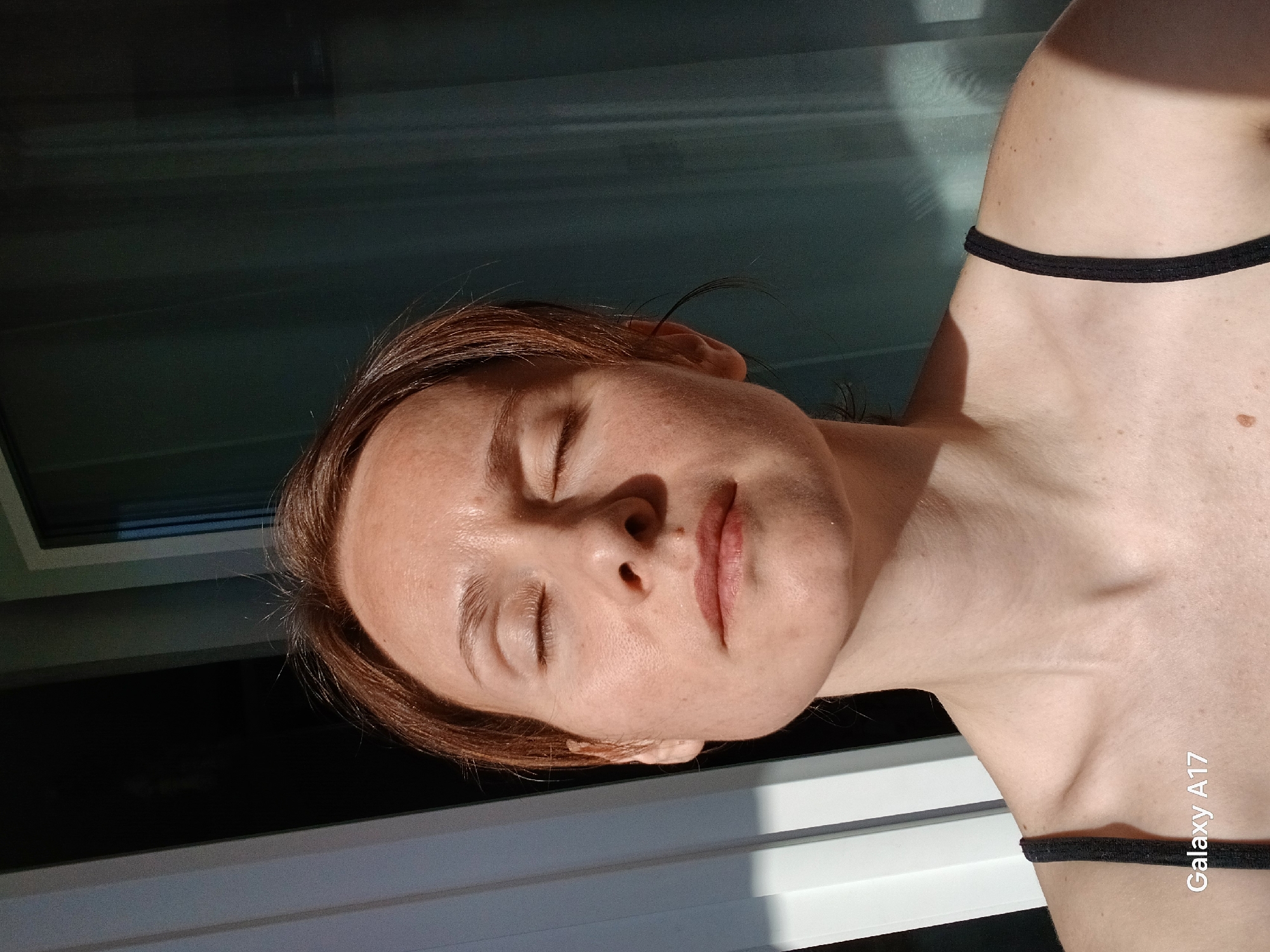 A photograph depicts a fair-skinned person with reddish-brown hair and closed eyes. The person is wearing a black-strapped garment or undergarment. The skin exhibits some freckles or moles. The background features a dark, possibly reflective surface with linear light patterns. The photograph was taken with a Galaxy A17. The location cannot be determined from the image.