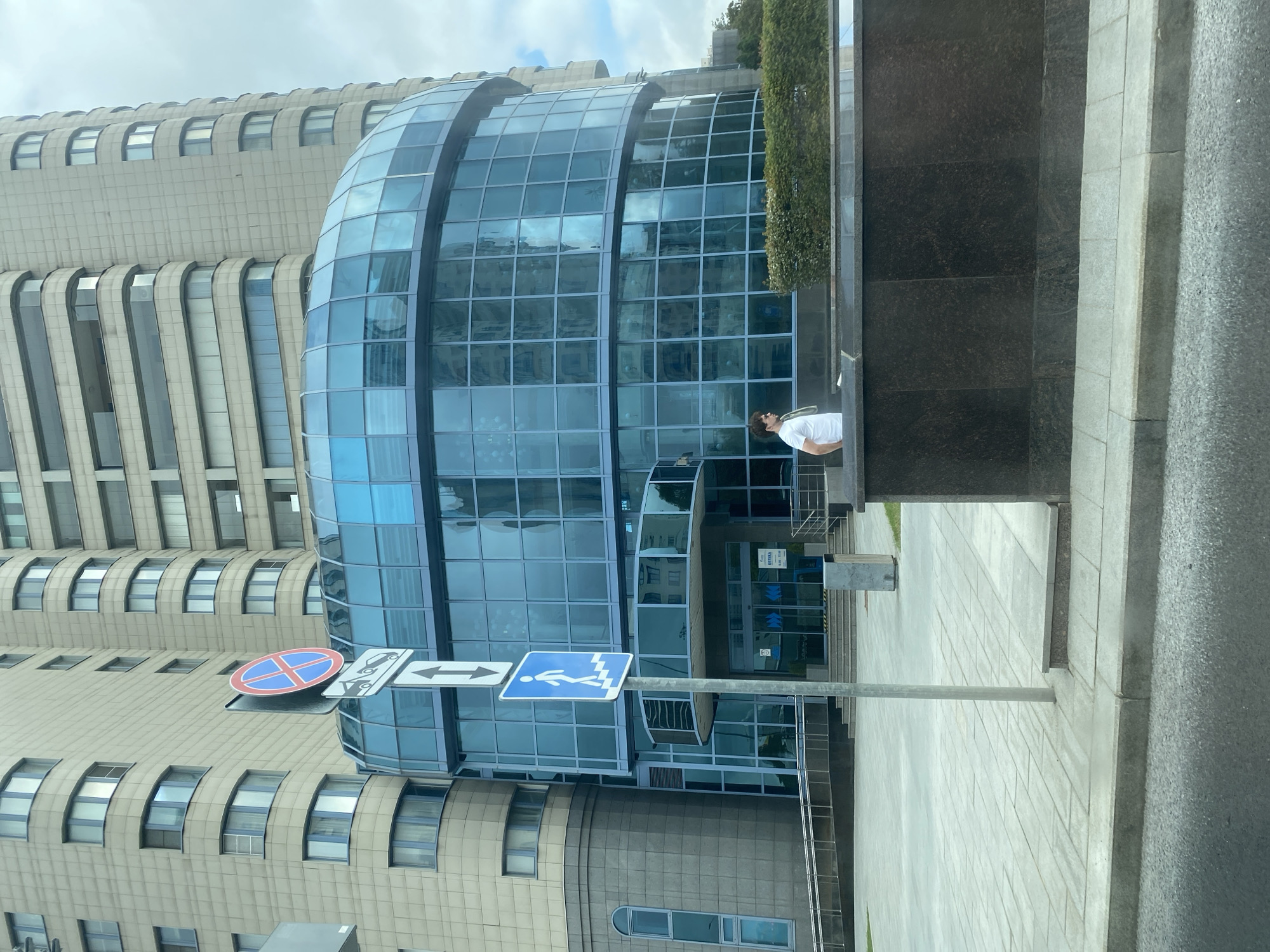The image shows a modern building with a curved glass facade, located in Moscow, Russia. In the foreground, a man in a white t-shirt stands near a low stone wall with a manicured hedge behind it. A street sign with pedestrian and no-parking symbols is visible on the left. The building behind him is a multi-story structure with tan-colored cladding and numerous windows. The sky is overcast.