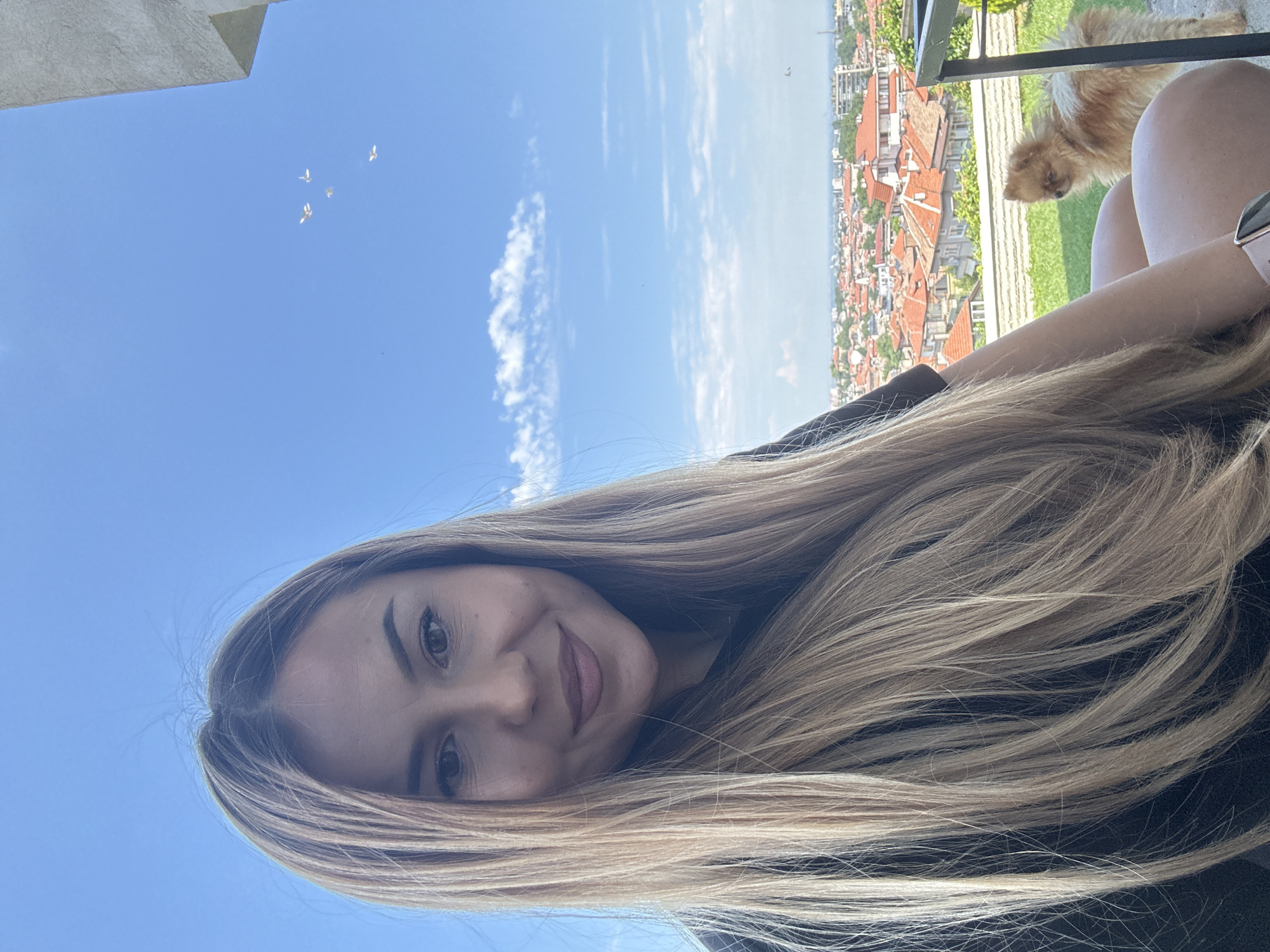 A young adult female is positioned in the foreground, looking directly at the camera with a slight smile. She has long, blonde hair with darker roots, parted slightly off-center, and visible flyaway hairs at the crown. She wears a dark-colored top, and a rose gold smartwatch is visible on her left wrist.
To her right and slightly behind, a small, light brown, fluffy dog, resembling a Pomeranian, stands on green grass, facing away from the camera and looking towards the distant city.
The background presents a panoramic view of the city of Asenovgrad, Bulgaria, observed from an elevated vantage point. The cityscape is predominantly composed of buildings with distinct red-tiled roofs, interspersed with green trees and vegetation. A textured, light-colored retaining wall spans the middle ground, separating the immediate grassy area from the urban sprawl. A dark metal railing is partially visible on the far right.
The sky above is clear and bright blue, featuring scattered white cumulus clouds. In the upper right quadrant, five small, white, bird-like objects are in flight.