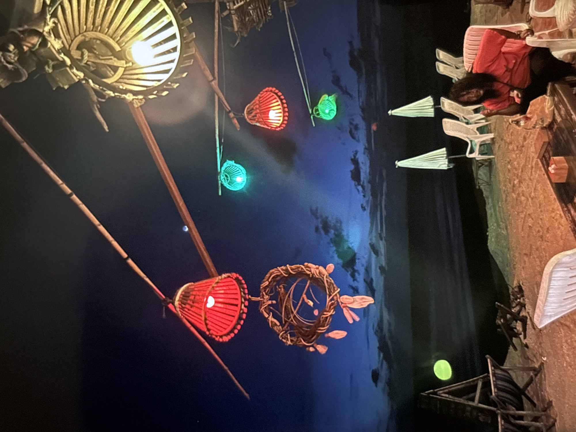 In the city of Chalong, Thailand, an outdoor nighttime scene unfolds on a sandy beach. Overhead, bamboo structures support decorative lights. A prominent red lantern, made of woven material, hangs prominently, accompanied by a circular woven decoration below it. Other lanterns, colored green and red, are also visible. In the foreground, a person wearing a red shirt is seated on a white chair, looking towards the viewer. A table with a box and a bag sits in front of them. Beach chairs and white umbrellas are set up on the sand, with the ocean visible in the background under a twilight sky. The overall ambiance suggests a beachside establishment open for evening patrons.