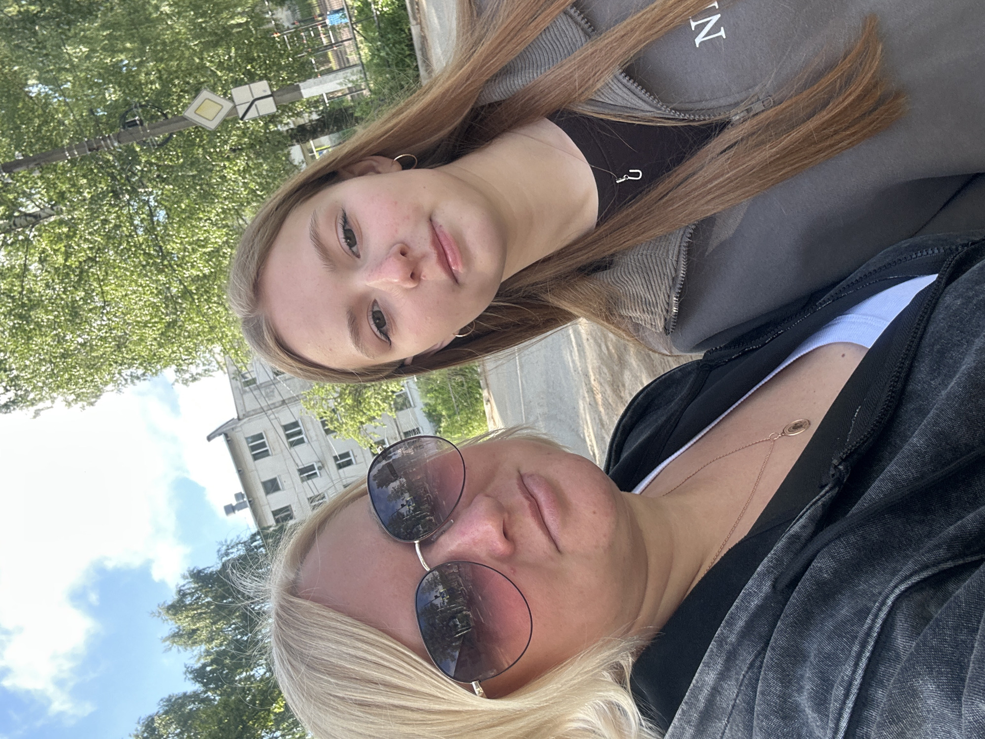 Two individuals, an adult female and a juvenile female, are taking a selfie outdoors in an urban environment. The adult female, on the left, has blonde hair and is wearing large, dark-rimmed sunglasses, a black hooded jacket, and a white tank top with a gold necklace. The juvenile female, on the right, has long, light brown hair and is wearing hoop earrings, a dark t-shirt with a silver necklace featuring a "U" charm, and a grey zip-up jacket.
The background consists of a multi-story apartment building, trees, a street, and a utility pole with traffic signs. The building appears to be a typical Soviet-era residential block. The overall setting suggests a residential area in the city of Chaikovsky, Russia, likely during daylight hours. The sky is partly cloudy. The image quality is consistent with a handheld smartphone photograph.