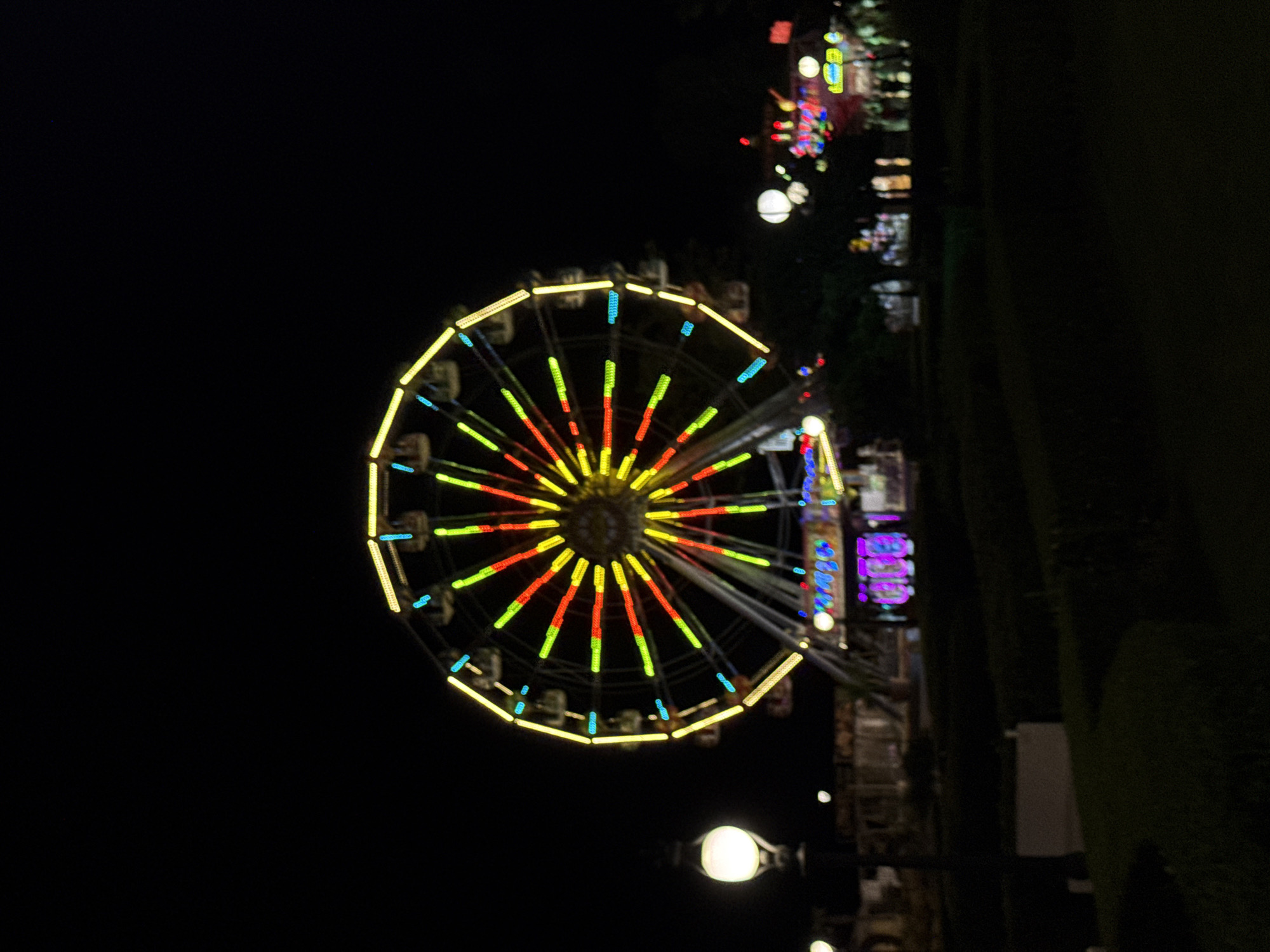 A Ferris wheel is illuminated with colorful lights against a dark night sky, likely in Varna, Bulgaria. The wheel's spokes are outlined in yellow, with red, green, and blue lights adorning the radial bars and car enclosures. In the foreground, a pathway with a black lamppost on the left is visible, along with manicured bushes. The background shows more illuminated structures and trees. The scene appears to be at an amusement park or fairground.