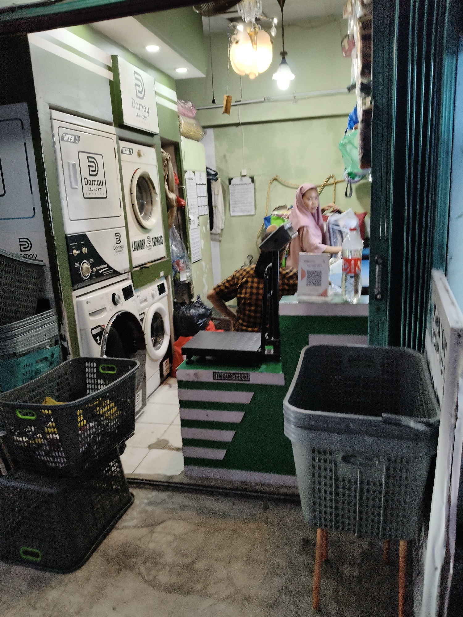 A laundry shop in South Tangerang, Indonesia, is depicted in this image. The shop is equipped with several washing machines and dryers, some of which have the "Damay Laundry Express" logo. A woman wearing a pink headscarf is seen operating behind the counter. She appears to be attending to a customer or managing operations within the shop. A QR code is visible on the counter, likely for payment or information purposes. Laundry baskets, both empty and containing clothes, are stacked or placed around the shop. The interior is illuminated by artificial lights, and the entrance is covered by a green metal gate. The overall scene suggests a functional and busy laundry service establishment.