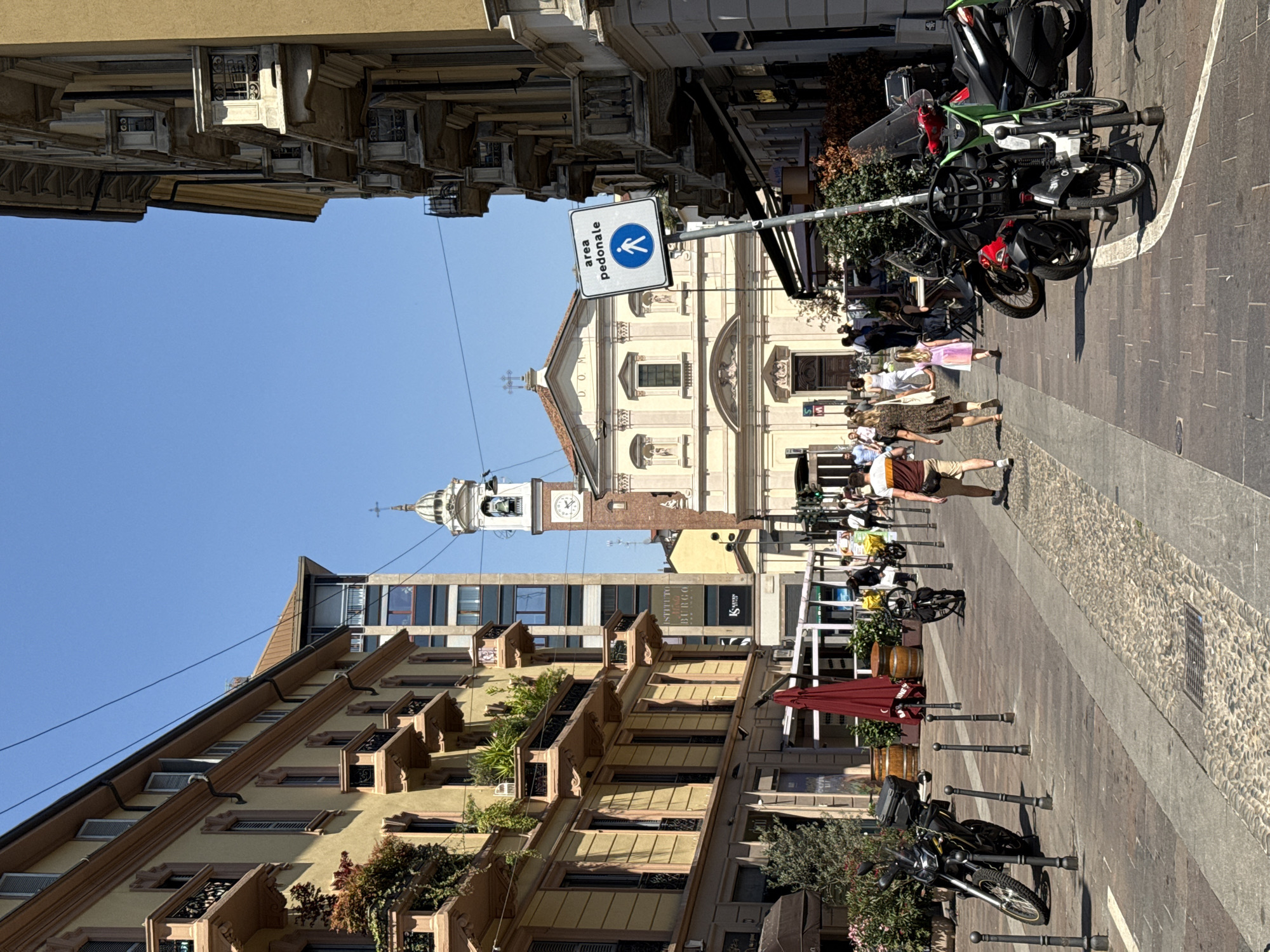 The image is a street scene in Milan, Italy. It is a sunny day with a clear blue sky. The view is down a narrow cobblestone street lined with buildings on either side. The buildings are tall, with ornate facades and balconies. The street is crowded with pedestrians, bicycles, and motorcycles.
In the center of the image is the Chiesa di San Sebastiano, a church with a cream-colored facade and a tall bell tower. In front of the church is a pedestrian sign.
The people in the image are dressed casually and appear to be tourists or locals going about their daily business. Some are walking, others are standing and talking. Several motorcycles and scooters are parked along the side of the road. There are also small shops and cafes visible in the background, adding to the bustling atmosphere of the scene.
