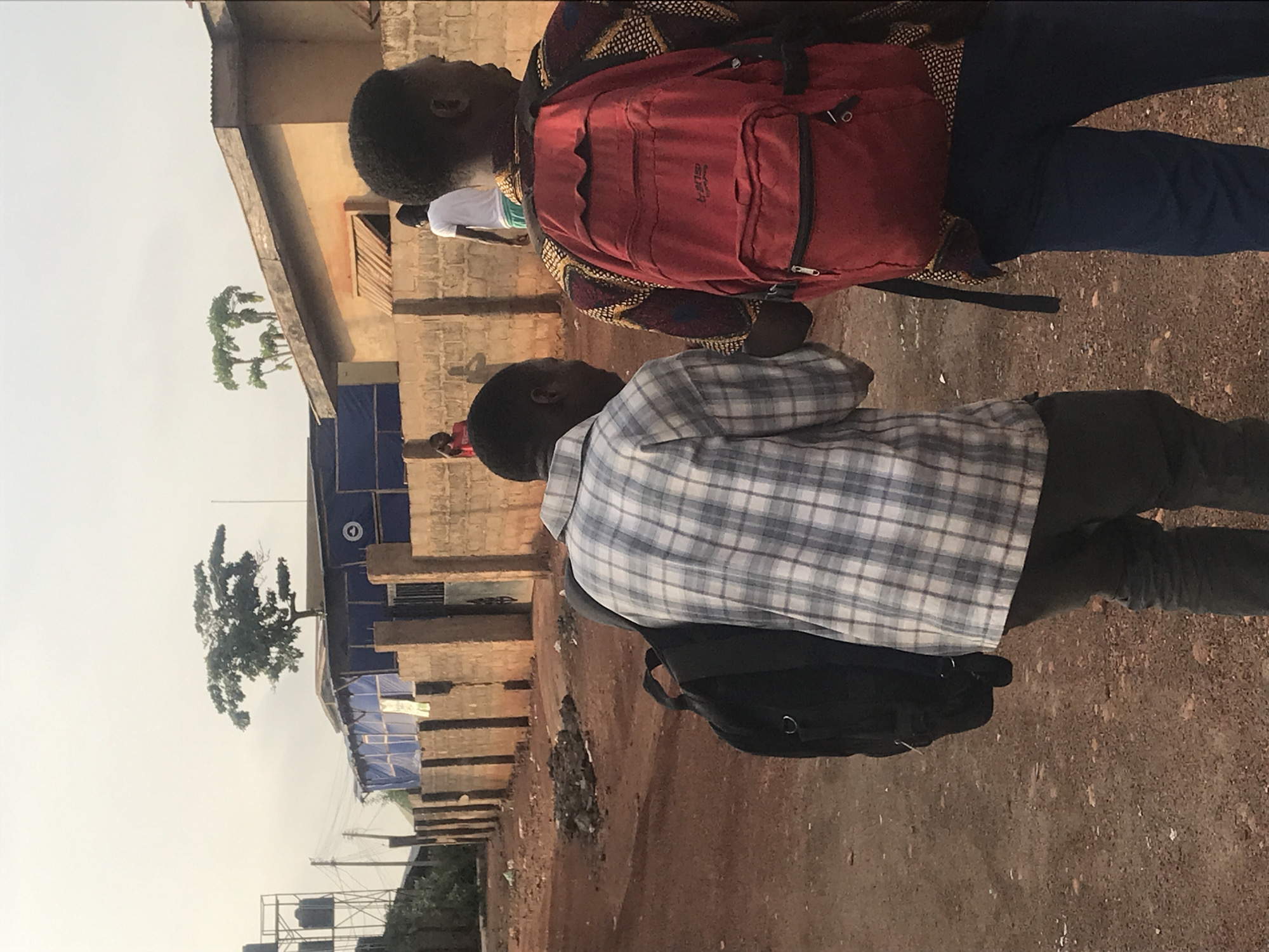 Two young men are walking along a reddish-brown dirt road next to low concrete buildings and a tree in Ilorin, Nigeria. One wears a red backpack, patterned shirt, and blue pants. The other wears a grey plaid shirt, dark pants, and a black backpack.