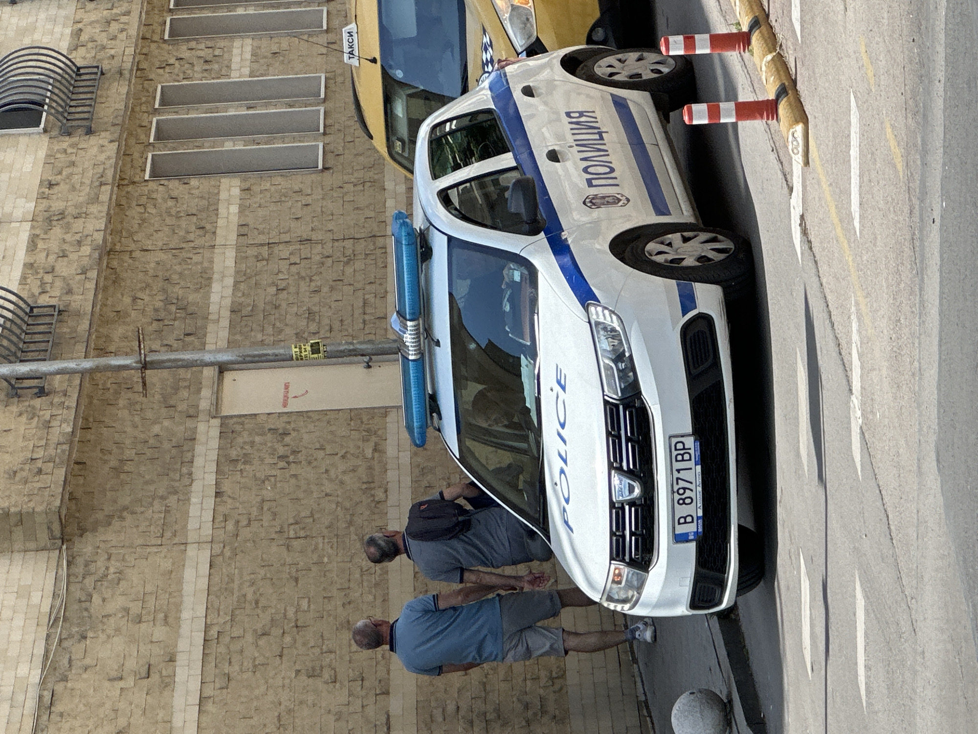 Two men stand near a white Dacia police car with the license plate B 8971 BP on a street in Varna, Bulgaria. A yellow taxi is parked behind the police vehicle. The scene is set against a brick building with rectangular windows and decorative metalwork. Traffic cones are visible on the side of the road. The image appears to be taken during daylight.