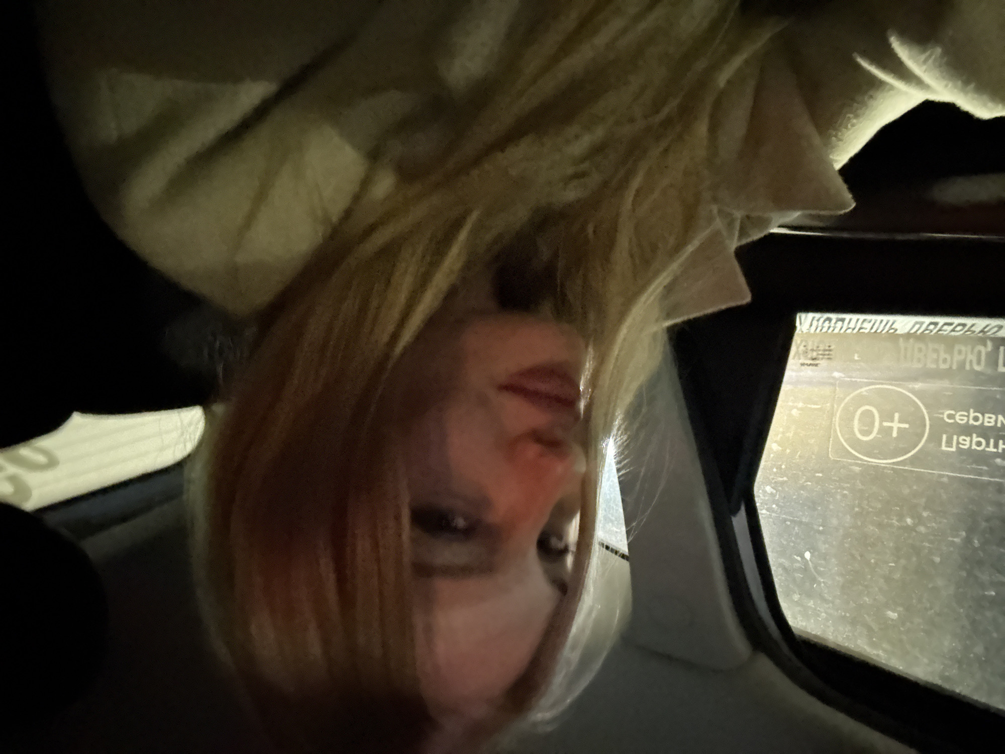 A young woman with blonde hair and fair skin is pictured inside a vehicle at night, with the city of Perm, Russia, suggested by Cyrillic text on a window sticker. The woman is looking towards the camera with a slight smile, her lips parted. The interior of the car is dimly lit, with some reflections visible on the window. The woman is wearing a light-colored jacket. The overall scene appears to be a candid shot taken during a car ride.