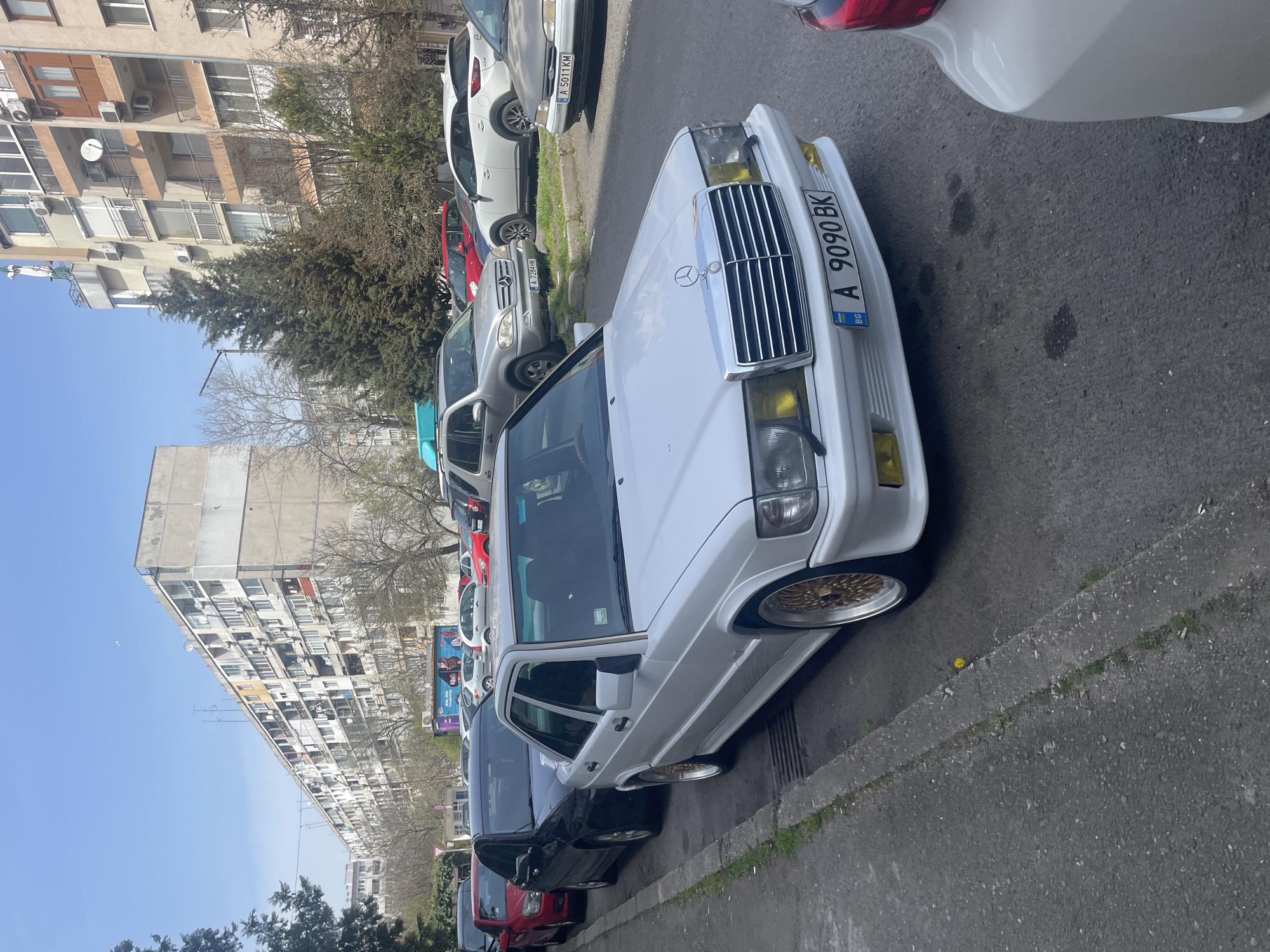 The image shows a street in a city, likely Burgas, Bulgaria, based on the license plate. A white Mercedes-Benz is parked on the right side of the street. A series of other cars are parked behind it, including gray, red, and black vehicles. Buildings, mainly apartment blocks, line the left side of the street. The weather appears sunny. The street is paved. There are trees along the side of the road. The car's license plate reads "A 9090 BK". The front lights of the Mercedes are yellow.