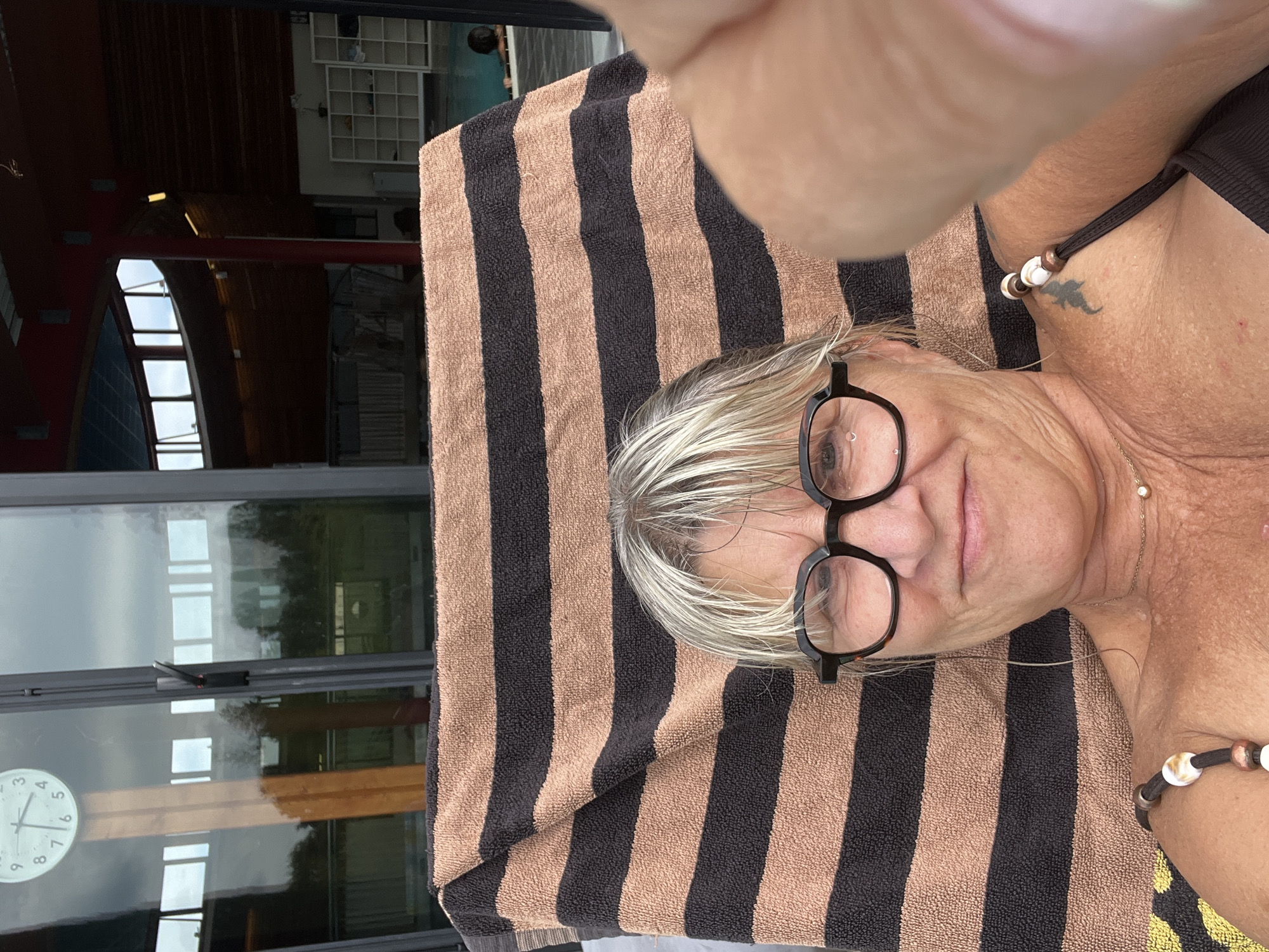 The image shows a woman lying on a striped towel in an indoor swimming facility. She is wearing glasses and a dark swimsuit. The background reveals a pool area with other people, a clock on the wall, and large windows. The setting appears to be the Casino de Montrond-les-Bains' thermal spa in France, which offers swimming and relaxation facilities.
