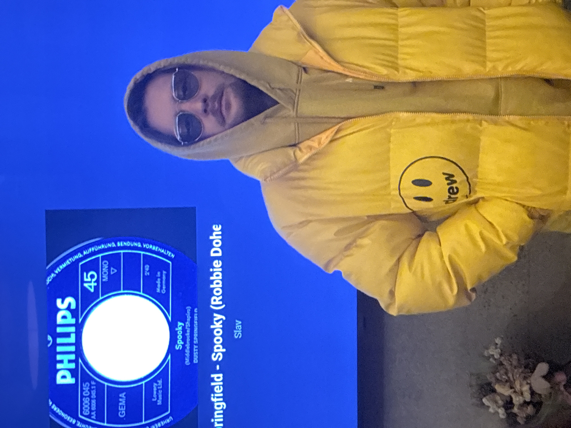 A man wearing a yellow puffer jacket and sunglasses stands in front of a television displaying an image of the record label for "Spooky" by Dusty Springfield. The jacket has a "drew" logo on it. The background behind the man and the television are not clear, although some floral decorations are visible. The exact location in Huixquilucan de Degollado, Mexico, cannot be determined from the image.