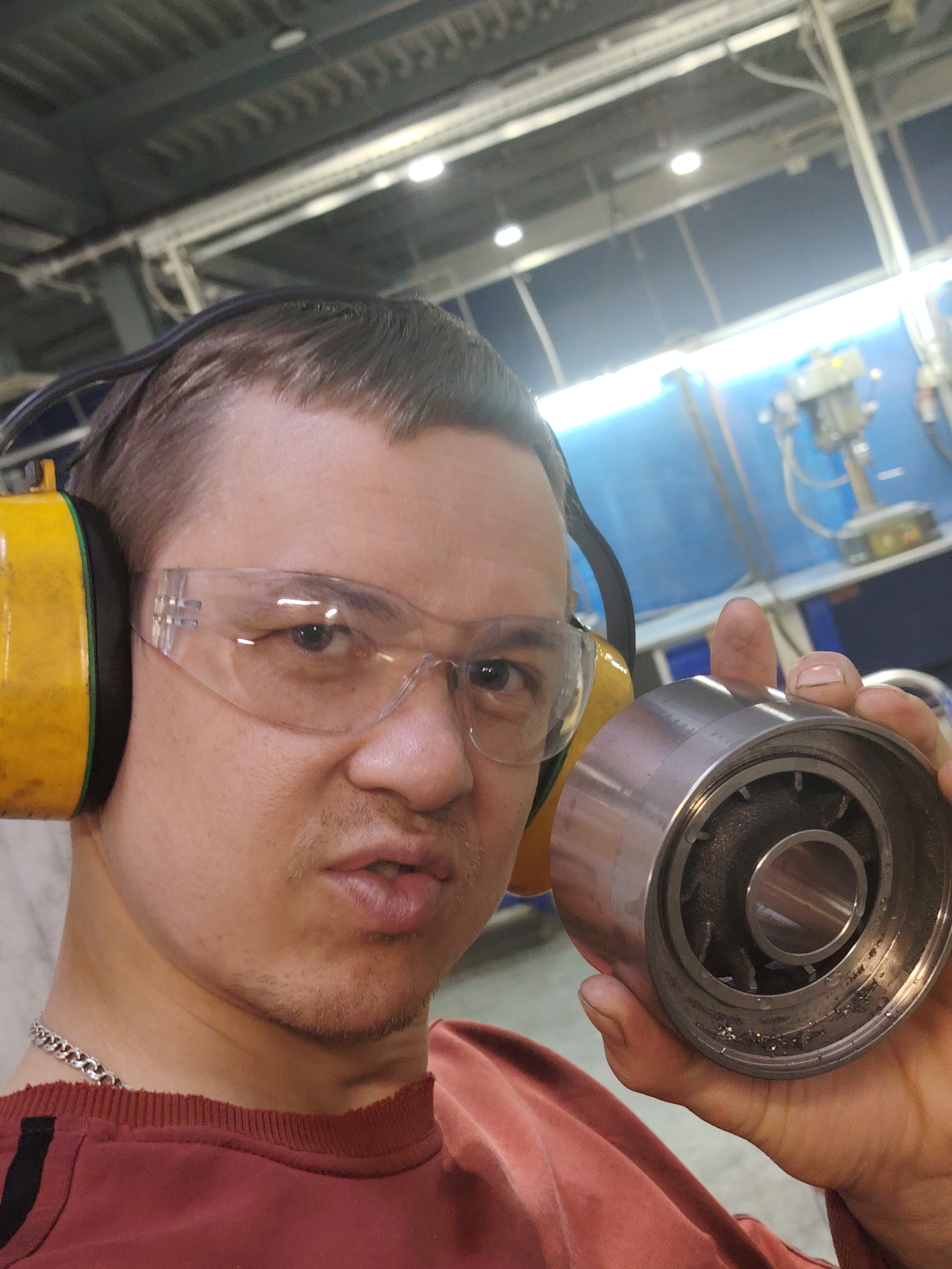 A man, likely in his 20s or 30s, is featured in a close-up, waist-up selfie, looking directly at the camera with a focused expression and slightly pursed lips. He is wearing clear safety glasses and bright yellow ear defenders, indicating a noisy or industrial work environment. His attire includes a reddish-brown long-sleeved shirt and a silver chain necklace. His short, dark hair is visible, along with light stubble on his chin and upper lip.
In his right hand, the man holds up a metallic, cylindrical machine component, possibly a bearing or roller. The object's interior reveals mechanical elements and a significant amount of small metallic debris, resembling swarf or fragments from wear, accumulated within its lower rim. This suggests the component may be damaged or has recently been removed from operation for inspection.
The setting is an indoor industrial facility, consistent with a workshop or factory in Noviy, Russia. The background features overhead industrial lighting and a ceiling constructed with metal beams. A prominent blue wall on the right side of the frame displays additional machinery, including what appears to be a drill press or similar equipment. The artificial lighting throughout the space is characteristic of an active manufacturing or repair site. No visible text is present within the image.
