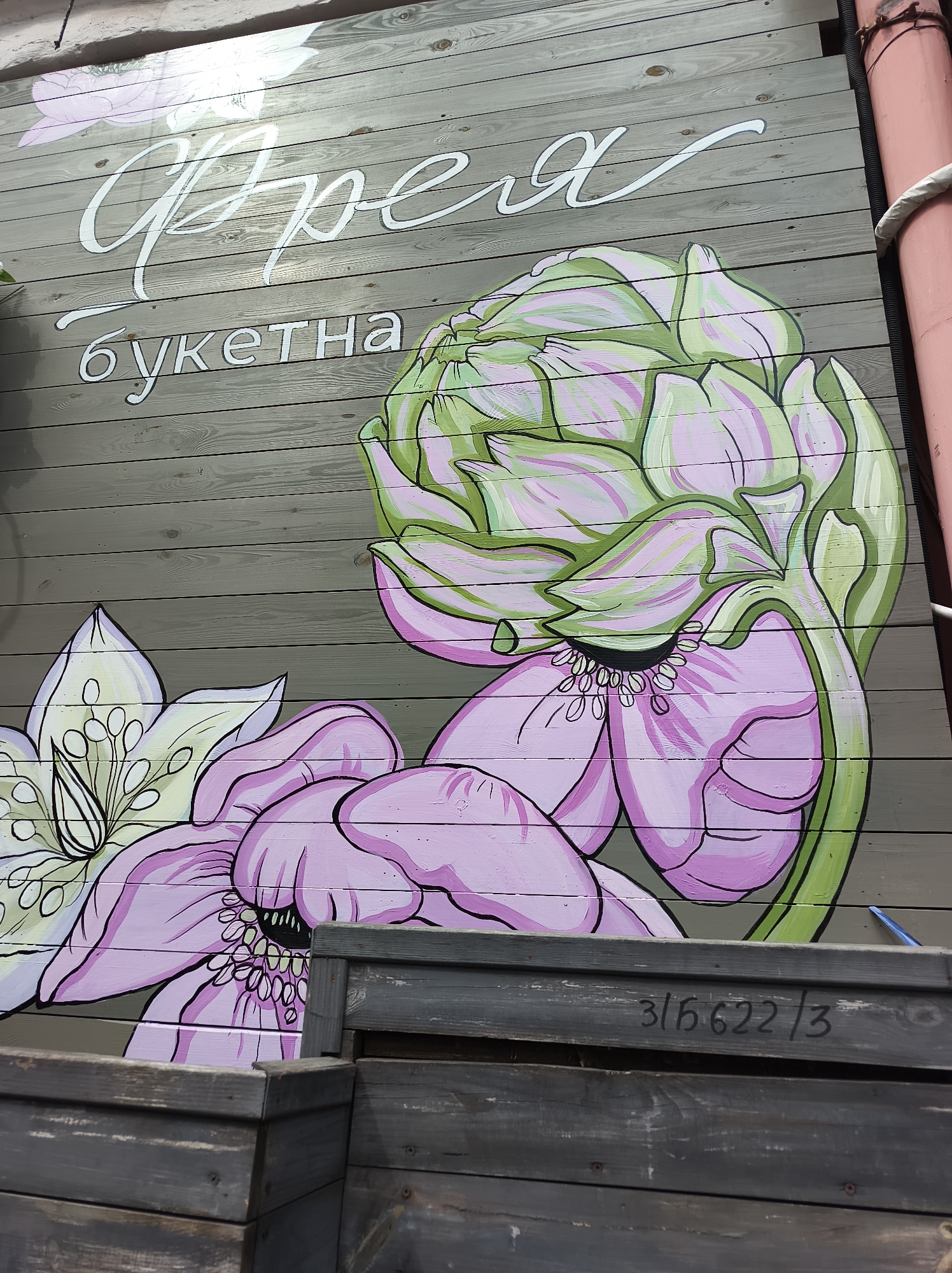 A mural, possibly advertising floral arrangements or a flower shop, is painted on a weathered wooden wall. The mural features stylized flowers, including a large artichoke flower and a flower with white petals and exposed stamens. Above the flowers are white, calligraphic-style words in Ukrainian, possibly the name of the shop, followed by the word "букетна" (bouquet). The wall is made of horizontal wooden planks with varying shades of gray. In the lower portion of the image, there are dark gray wooden boxes. A red pipe and a white rope are visible along the right edge of the wall. Writing in black paint, "3/56622/3", is visible on one of the wooden boxes near the bottom right. The mural likely is located in Kyiv, Ukraine, due to the presence of Ukrainian text.