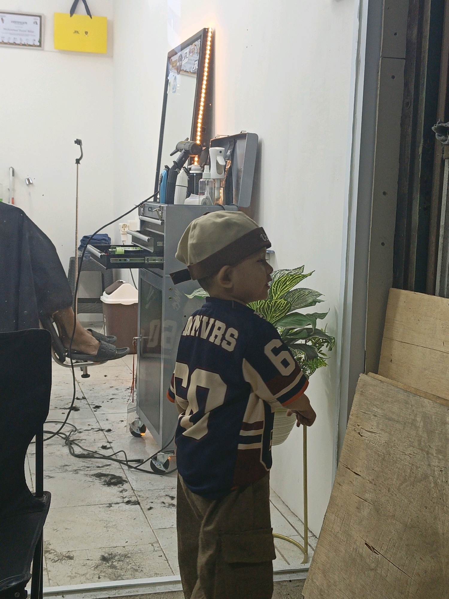 A young boy wearing a baseball cap and a dark blue jersey with "ANVRS" and the number "27" on it stands facing a wall in what appears to be a barbershop. He is looking towards a plant with large green leaves. The floor is tiled and scattered with hair clippings. To the left, a person in a black apron, likely a barber, is seated in a chair with their feet resting on a second chair. In the background, a mirror with LED lighting is visible, along with various barber tools and products on a trolley. A yellow shopping bag hangs on the wall in the upper left corner. The scene is likely located in the city of Pangkajene, Indonesia, given the context of previous images.