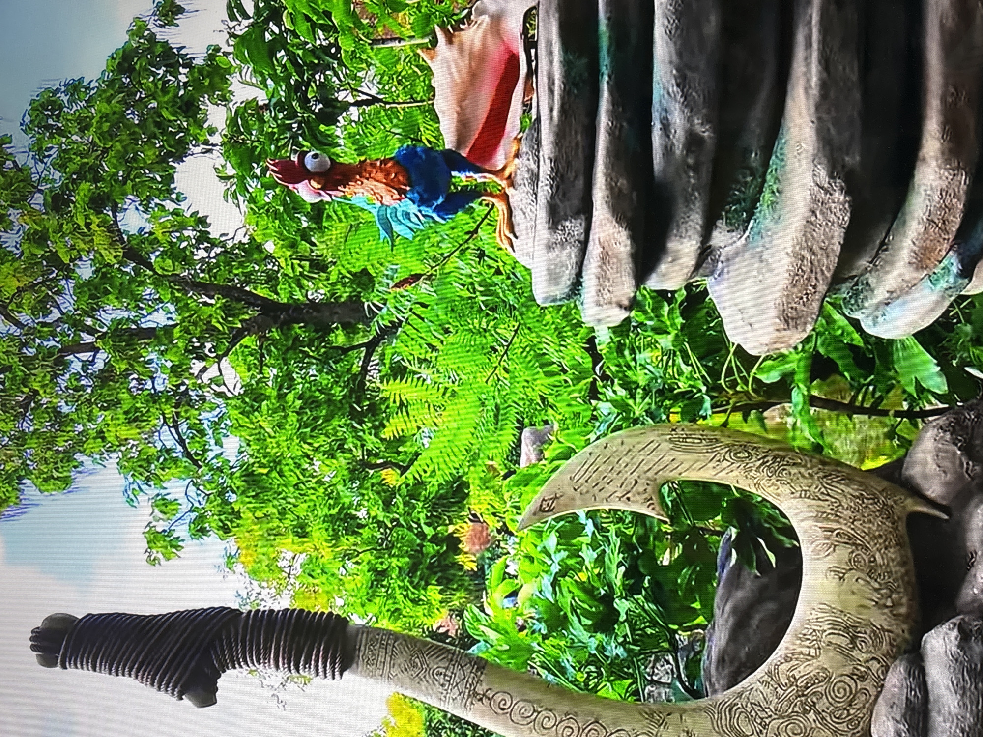 The image depicts a scene from the Moana Journey of Water attraction at Epcot in Walt Disney World, Florida. The objects visible include a figure of Hei Hei (a rooster character from the movie Moana), a section of a rock formation, and a portion of Maui's hook. The environment is dominated by lush greenery, simulating a tropical island landscape.