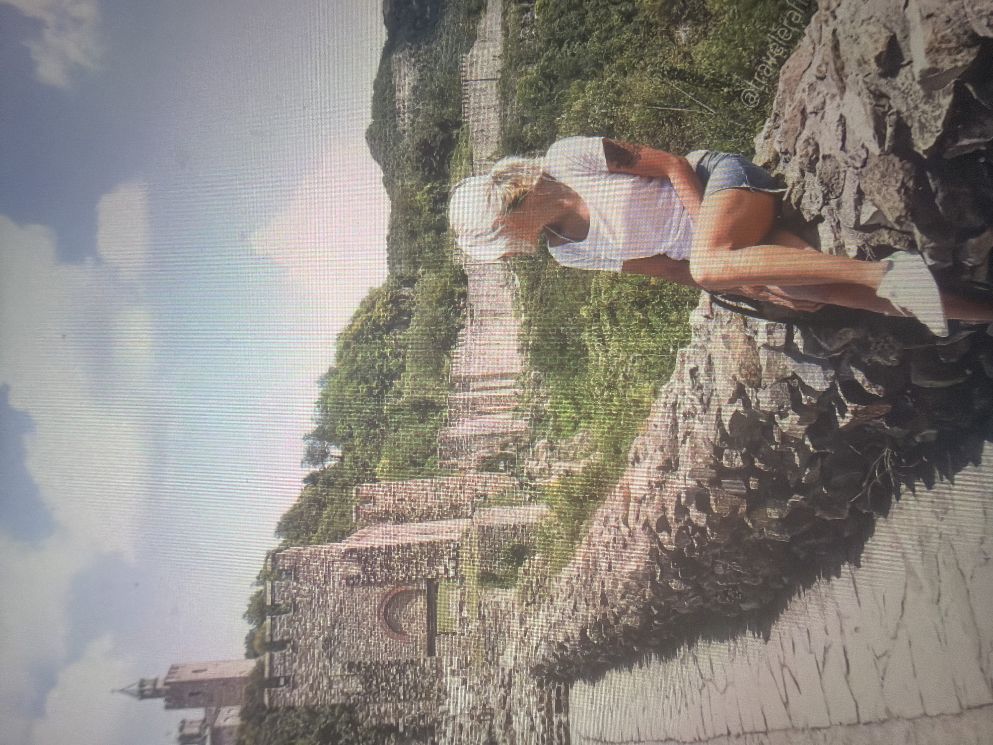 A woman with light-colored hair, wearing a white t-shirt, denim shorts, and white sneakers, sits on a large stone formation in the foreground on the right side of the image. A tattoo is visible on her right upper arm. She is positioned facing towards the left of the frame. In the mid-ground and background, an extensive stone fortress complex is visible, situated on tree-covered hills under a partly cloudy sky in the city of Shumen, Bulgaria. To the far left, a multi-story stone tower with a peaked roof and arched windows stands on a hilltop. Below this tower, a long section of stone walls with battlements and an arched gateway extends across the landscape. Further to the right, more segments of ancient stone walls are visible winding along the hillsides. A paved pathway runs along the left side of the foreground, bordered by a low stone retaining wall.