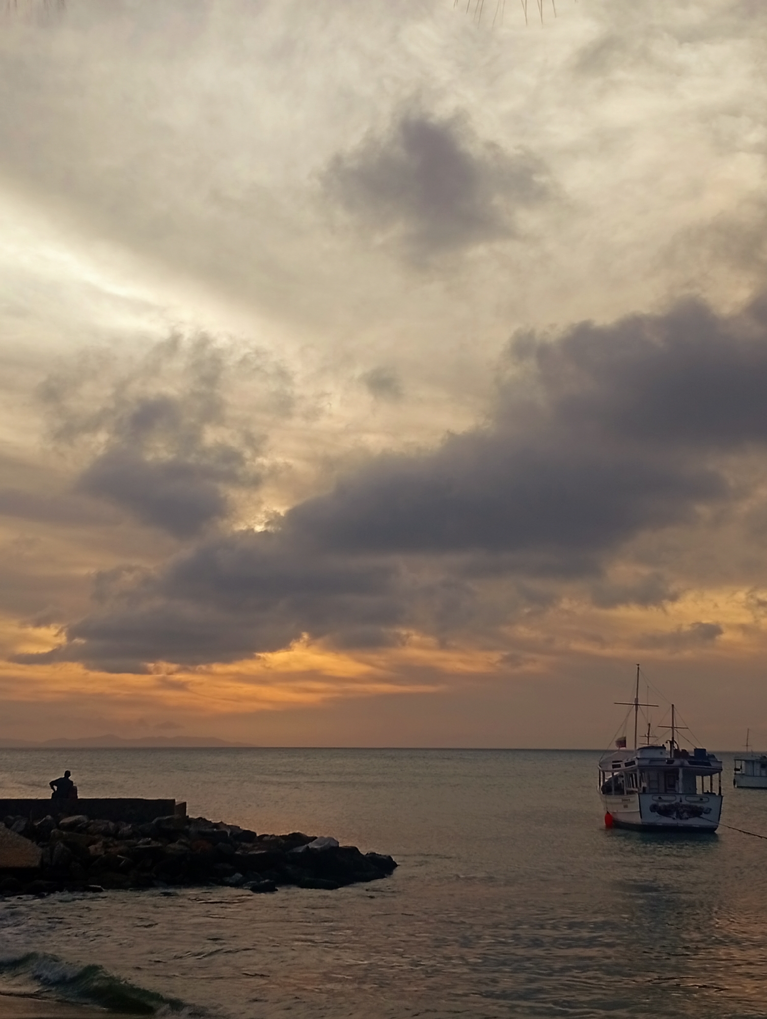 The image displays a coastal scene in Juan Griego, Venezuela, during sunset. The sky, occupying the upper two-thirds of the frame, features a mix of dark grey stratocumulus clouds and lighter, diffused altostratus clouds, interspersed with areas of bright orange and yellow light from the low sun, particularly towards the horizon. The water below reflects these colors, appearing dark blue-grey with warm undertones. Small waves are visible breaking near the foreground shore on the left.
On the left side of the frame, a dark, rocky breakwater extends into the sea, topped by a low, dark concrete structure. A single, silhouetted individual is seated on this concrete structure, facing towards the right side of the frame and the horizon. In the mid-ground on the right, a white fishing or pleasure boat with a cabin, mast, and rigging is anchored in the water. A red buoy or fender hangs from its port side, and dark, possibly painted, details are visible on its bow. A portion of a second, similar boat is faintly visible further to the right. A faint outline of distant landmasses is discernible on the horizon to the left of the main boat.
