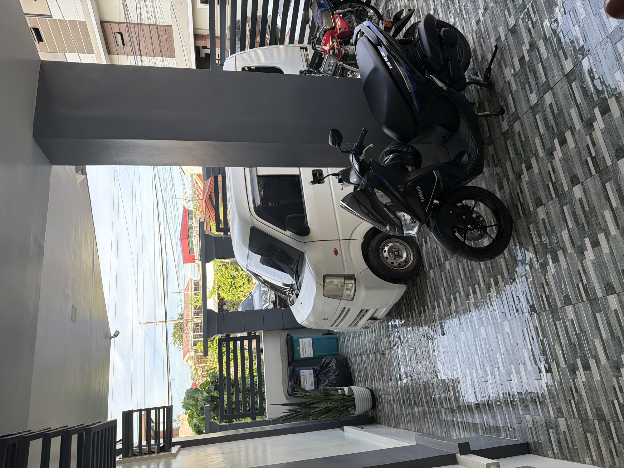 The image shows a carport in Davao City, Philippines. A white van and two motorcycles are parked on a gray-tiled surface. There is a small garden area behind a black metal fence, with trash bins and a potted plant visible. Overhead, there are power lines against a blue sky. The buildings in the background appear to be residential houses. The scene is taken during the daytime, with natural lighting.