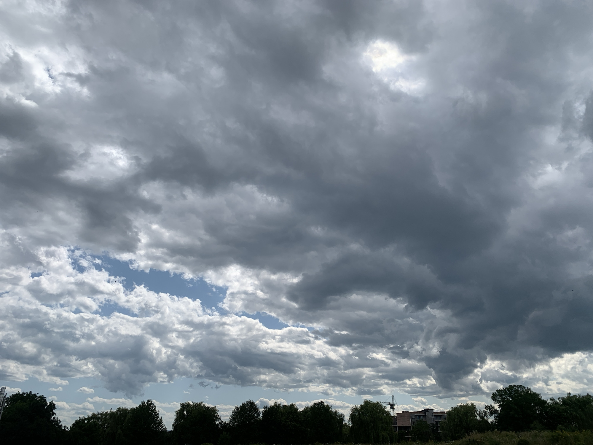 The image depicts a cloudy sky over a landscape. The clouds are varied, with some appearing dark and heavy, suggesting impending rain, while others are lighter and show patches of blue sky. The lower portion of the image features a dense line of trees silhouetted against the sky, with some buildings and a construction crane visible in the distance. The scene appears to be a natural or semi-urban environment. The specific location in Kolomyia, Ukraine is not discernible from the visual information provided in the image. There are no people or discernible actions taking place.