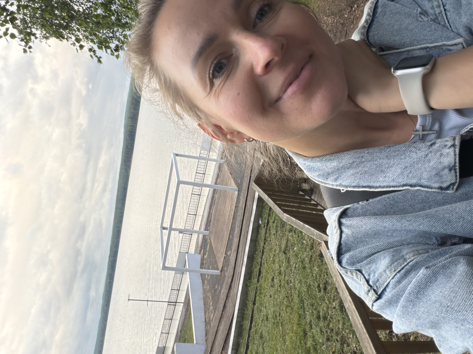 A selfie-style portrait depicts a Caucasian woman in an outdoor setting within the city of Chaikovsky, Russia. The woman, estimated to be in her 20s or 30s, occupies the lower right quadrant of the image, facing the camera with a subtle smile and visible periorbital rhytides (crow's feet). Her hair is light blonde/brown, styled in a casual manner with some strands loose around her face. She wears a light blue denim jacket over a light-colored, possibly striped, top. A silver chain with a small, unadorned cross pendant is visible on her neck. Her left wrist displays a light grey/beige Apple Watch with a silver case, and her left hand rests against her jawline.
The background features a vast body of water, likely a river or reservoir, stretching horizontally across the midground. The distant shoreline is marked by a continuous, dense forest treeline. Immediately behind the woman, a wooden railing suggests a viewing platform or descending path. Below this, a grassy slope transitions into a section of wooden planks or sleepers resembling tracks, running alongside a white concrete retaining wall. Further towards the water, a paved walkway with a black metal fence borders the water's edge. A white, open-frame structure, possibly a modern pergola, stands on a wooden platform by the water. A tall, dark, slender pole is also visible near the water. The sky is largely overcast with diffuse grey and white clouds. Green tree foliage is present in the upper right corner of the frame.