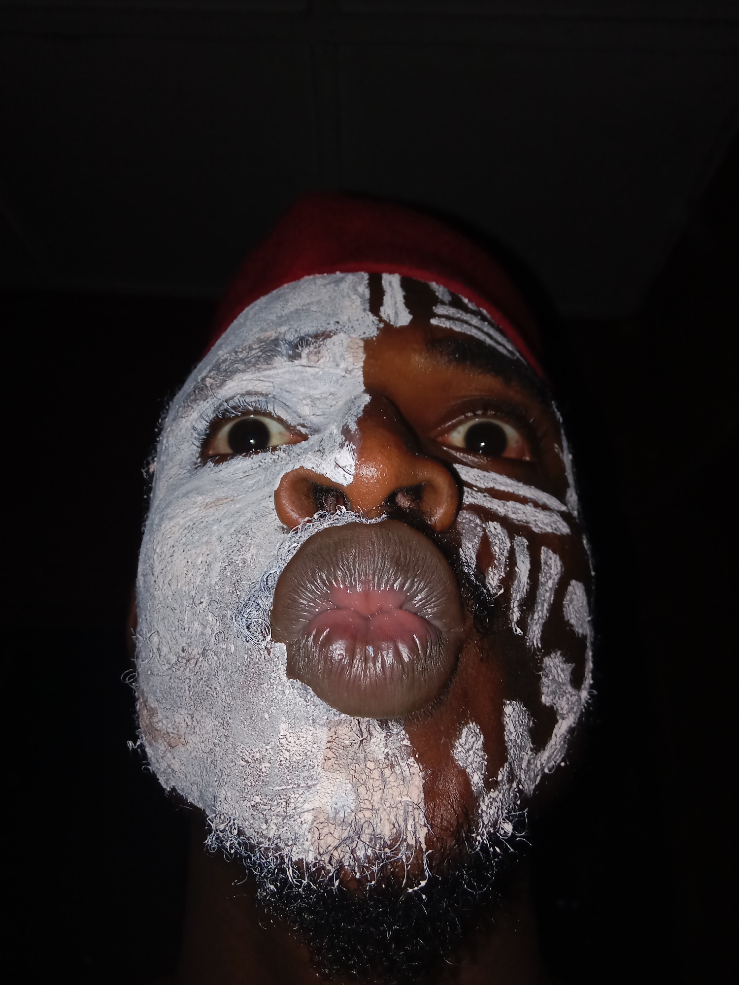 A low-angle, close-up portrait shot of a person with dark skin, wearing a red hat with white trim. The person's face is half-painted with white paint. The left side of the face is completely covered in white paint, while the right side is painted with tribal-like markings. The background is black. The person is puckering their lips and staring directly at the camera. The location is unknown.
