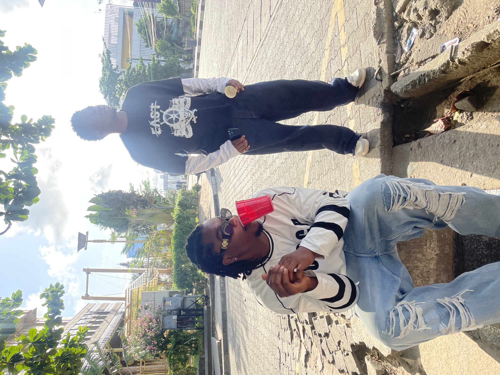Two young men are outside. One is standing, wearing a long-sleeved black shirt with a white design, and dark pants. He is holding a cup and a cell phone. The other is seated, wearing a long-sleeved white shirt with black stripes and distressed jeans. He is holding a red cup to his mouth. They appear to be on a city street, with buildings and greenery visible in the background.