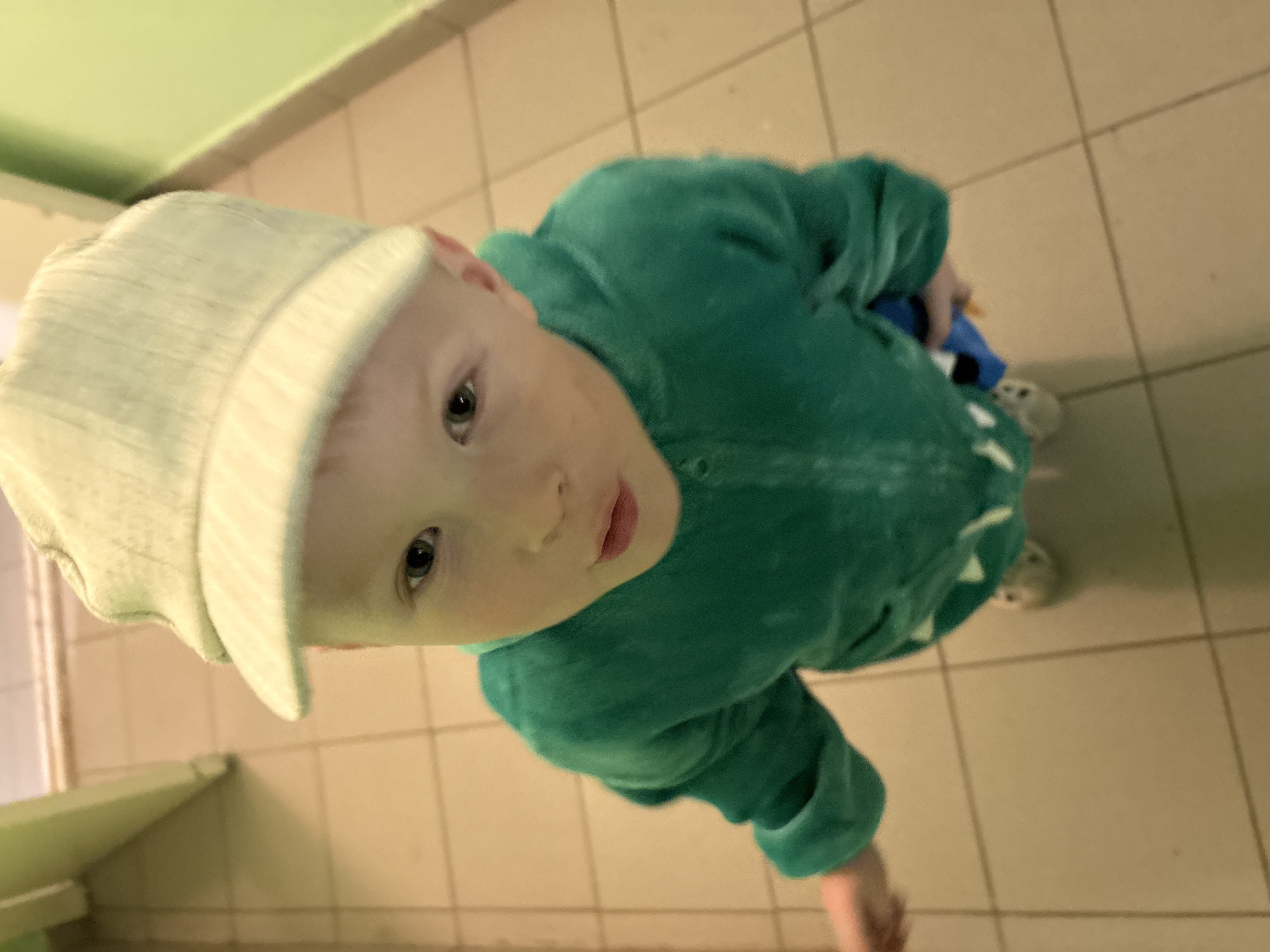 A young Caucasian boy, appearing to be around 3-4 years old, stands indoors, possibly in a hallway or corridor. He is wearing a light-colored baseball cap and a bright green hooded jumpsuit with a dinosaur-like design on the front. The boy's face is turned towards the camera, and he has fair skin, light hair, and blue eyes. He is looking slightly upward. The floor is tiled in a beige or cream color, and the walls are painted a pale green. The lighting is somewhat dim, casting subtle shadows. The image does not provide any specific information about the location within Izhevsk, Russia, or any particular actions or interactions occurring beyond the boy's presence and gaze.