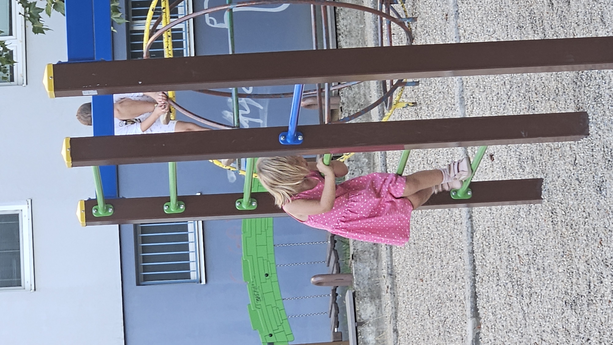 A young girl with blonde hair, wearing a pink polka dot dress and light pink shoes, is climbing on a monkey bar structure in a playground. She is gripping the green bars with her hands and her feet are placed on a lower green bar. In the background, another child, partially obscured, is visible, wearing a white shirt and shorts. The playground equipment includes brown wooden posts with yellow caps and green and blue bars. The ground is covered with light-colored gravel. Behind the playground, a blue building with barred windows and a white-framed window is visible. The scene is set in Bratislava, Slovakia, indicated by the graffiti on the building in the background.