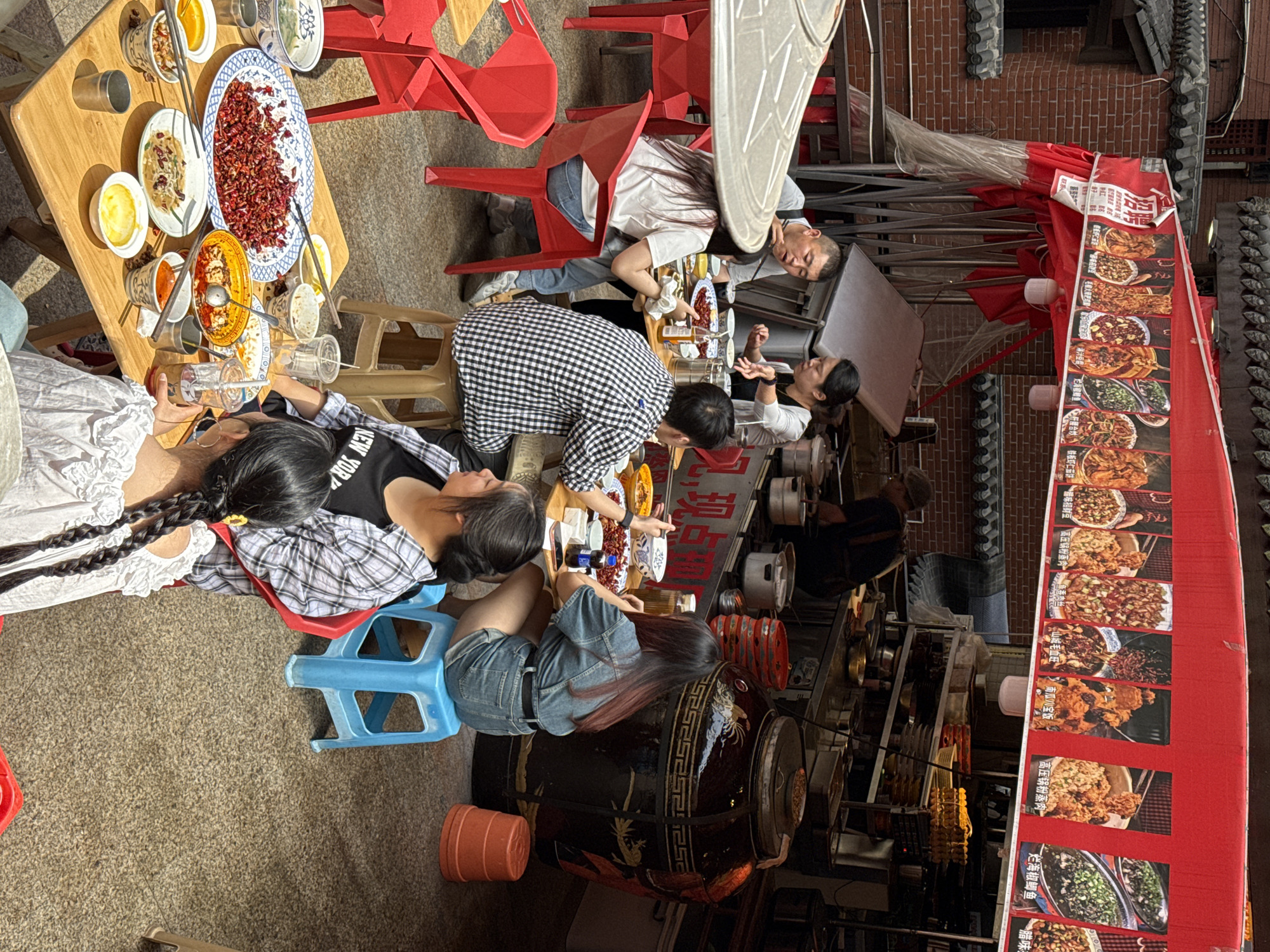 Here is a description of the image:
The photograph captures a bustling outdoor dining scene in what appears to be the Yuzhong District of China, based on architectural cues and the style of food signage. Several people are seated around a table laden with various dishes. The table is set with small bowls and shared plates, suggesting a communal dining experience. One person is wearing a black and white checkered shirt and bending over to look at the food.
A red awning with menu items displayed is overhead, showcasing a range of local cuisine. The menu features dishes such as "Iron Plate Shrimp with Eggplant Cake" and "High-Pressure Cooker Steamed Meat." The setting appears to be a street or alleyway with traditional brickwork visible in the background. A large, decorative jar or container is on the right side of the image, adding to the cultural atmosphere.