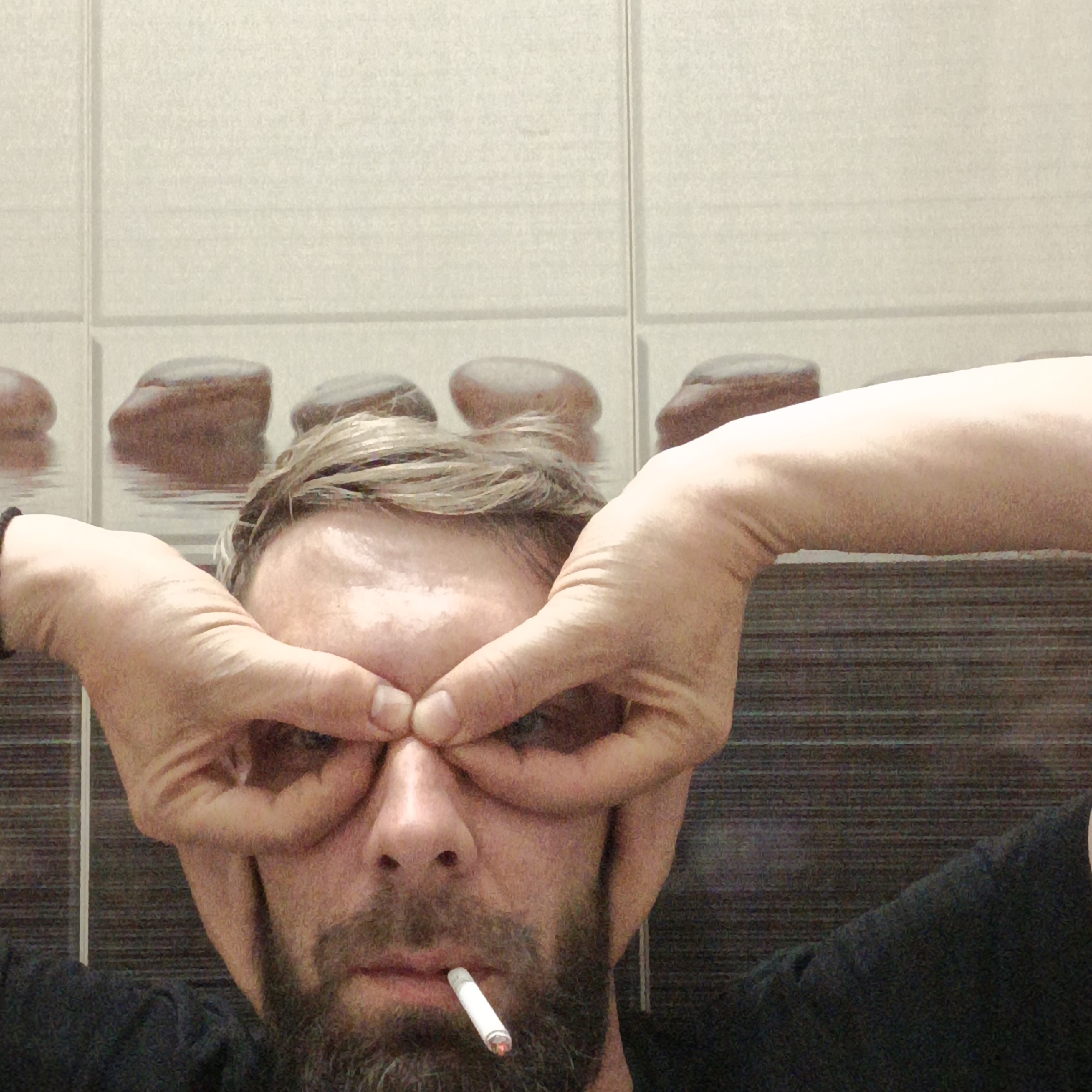 A man with a beard and a cigarette in his mouth makes binoculars with his hands in front of his eyes. He is indoors, standing in front of a wall covered in tiles. The location is Марковский, Russia.