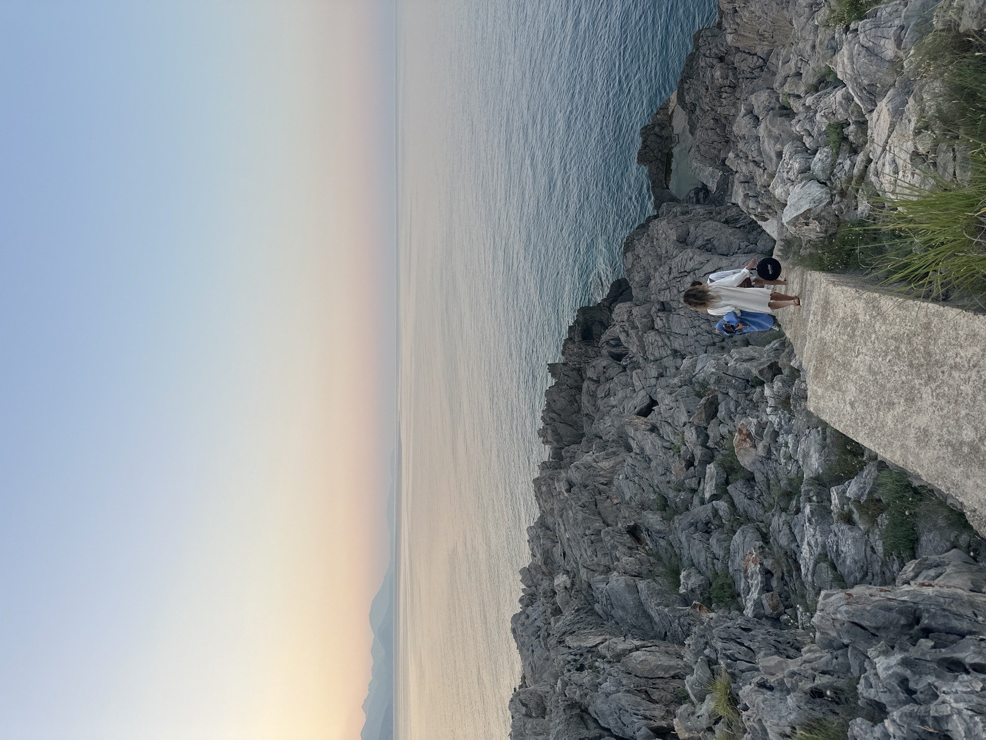 This image depicts a serene coastal scene at dusk in Dubraljevina, Montenegro. Two individuals are walking down a concrete staircase that winds through rugged, gray rock formations leading towards the sea. The person in front wears a white dress and carries a dark hat. The other person, slightly behind, is dressed in blue. The calm sea stretches to the horizon, where distant mountains are visible under a soft, gradient sky transitioning from pale pink and yellow near the horizon to a clear blue overhead. The rocky terrain is interspersed with sparse green vegetation, adding a touch of wildness to the landscape. The overall atmosphere is tranquil and picturesque, typical of the Montenegrin coastline during sunset.