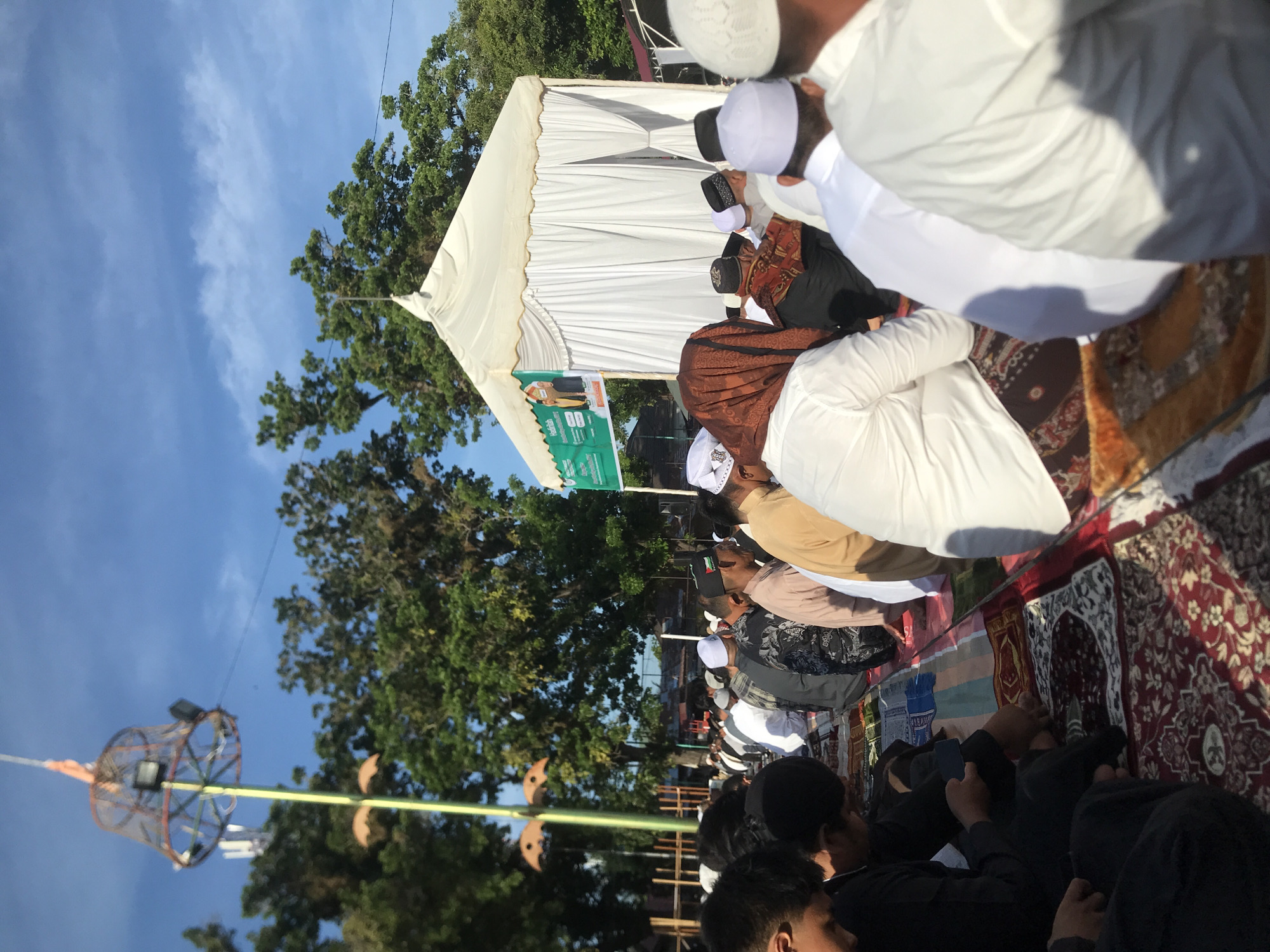 The image shows a group of men in Lhokseumawe, Indonesia, performing prayers outdoors. They are kneeling on prayer rugs, with some wearing white robes and caps. A white tent structure is visible in the background, along with trees and a blue sky. A decorative hanging structure is seen to the left.