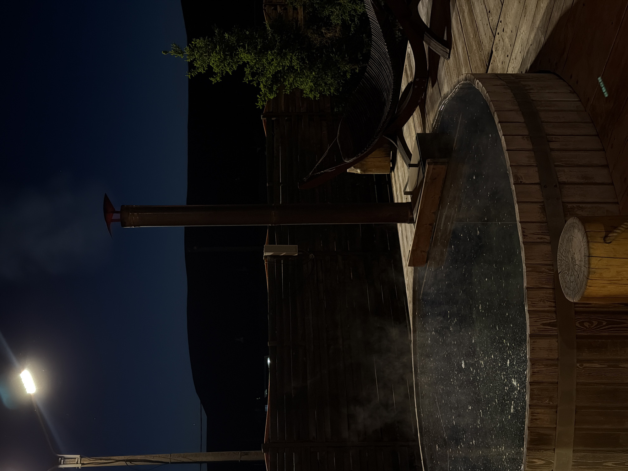 The image depicts an atmospheric outdoor scene at night, likely in Drokino, Russia. The central subject is a large, dark-colored hot tub or wooden barrel filled with water, from which steam gently rises, indicating warmth. The hot tub is encircled by metal bands, characteristic of traditional wooden barrels, and is set into or next to wooden decking or a platform.
Rising from near the hot tub is a tall, slender metallic pipe, topped with a distinctive red, fin-like element. This appears to be a chimney, possibly for a wood-fired heater that warms the water in the tub. Surrounding the area are various wooden structures, including a vertically planked wall or fence in the background. Green foliage from bushes or small trees is visible behind this wall, partially illuminated against the dark sky.
To the left of the chimney, a dark, curved, ribbed metallic structure is present, possibly part of a chair, decoration, or another architectural element. On the right side of the hot tub, a circular, intricately carved wooden disk is embedded into the surrounding wooden structure.
The scene is primarily lit by artificial sources, most notably a bright, rectangular outdoor light fixture mounted on a pole in the lower left of the frame, casting a strong glow and illuminating some parts of the scene while leaving the background sky a deep, dark blue. The overall ambiance is tranquil and inviting, suggesting a setting for relaxation. No people are visible, and there is no discernible text within the image.