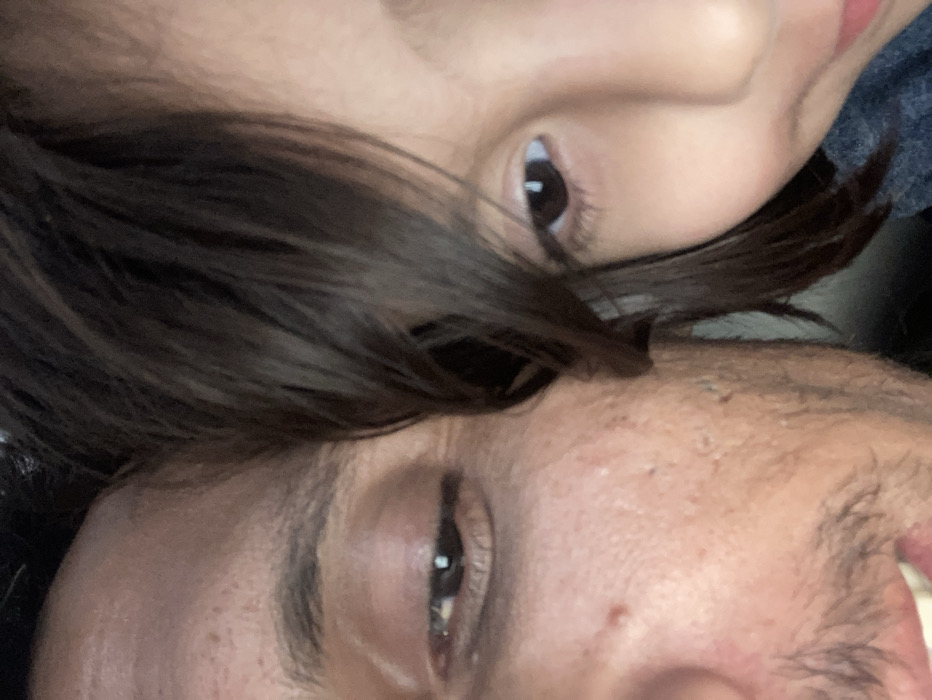 The image displays a close-up, vertical shot of two individuals' faces in extremely close proximity. On the left, the right side of an adult male's face is visible, including his forehead, a partially closed right eye with a visible dark iris, part of the bridge of his nose, and his lower cheek and jawline which show stubble. His skin exhibits some texture, and his mouth is slightly open, revealing upper teeth. His dark brown hair is visible on the top left. On the right, the left side of a young individual's face, consistent with a child, is visible. This includes a portion of their forehead, an open left eye with a dark iris directed slightly downwards and to the right, part of their nose, and a smooth cheek. Dark brown hair frames the child's face. A dark blue fabric is partially visible beneath the child's chin. The background is uniformly dark and featureless. The specific location within Quetta City Tehsil, Pakistan, cannot be determined from the visual evidence provided in the image.