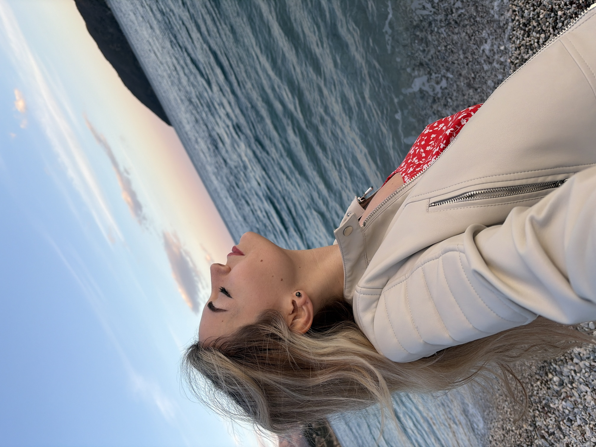 A blonde female individual is depicted in profile from the chest up, standing on a pebble beach in a coastal area of Budva, Montenegro, during what appears to be a sunset or sunrise. Her head is tilted backward, eyes closed, and lips are painted red. Her long, wavy blonde hair is wind-swept. She wears a cream-colored, quilted faux leather jacket with prominent zippers. A red garment with white floral patterns is visible beneath the jacket. A small black stud earring is present in her left ear. The environment includes turquoise-blue sea water with small waves breaking on the shoreline. A dark, prominent landmass, likely a hill or island, is visible in the mid-distance on the right. The sky overhead is light blue, transitioning to a pinkish-orange hue at the horizon, with sparse, wispy clouds.