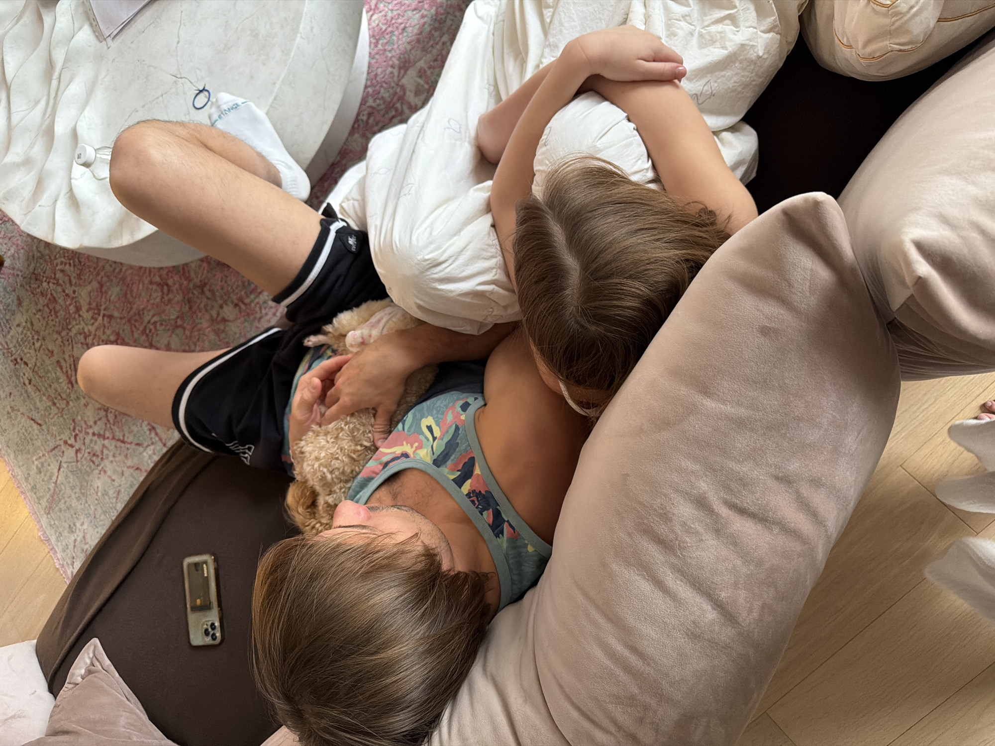 A high-angle shot captures a man and a child lying on a brown sofa amidst an assortment of pillows. The man wears black shorts with white stripes, a patterned tank top, and rests his head on a large beige pillow. A small, curly-haired dog lies between him and the child. The child, positioned with their back to the camera, is covered by a white blanket and rests against several pillows. Beside the man, on the sofa, is a cell phone with a case. Part of a round marble table and a patterned rug are visible in the background. The location cannot be determined to be Moscow, Russia based on the visual information provided.