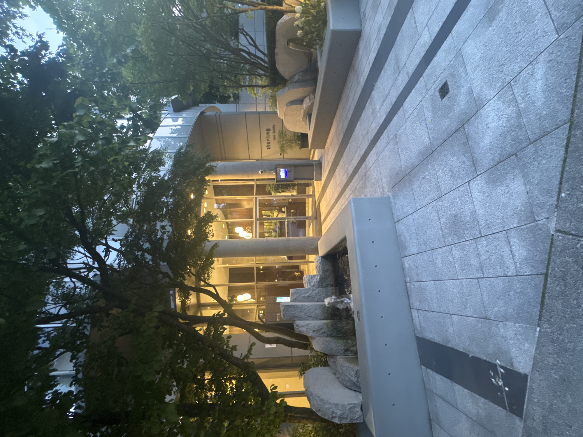 The image shows the exterior entrance to a building with the word "sterling" visible on its facade. The building appears to be a modern apartment or condominium complex. In front of the entrance, there is a paved walkway made of large, grey square tiles with darker grey rectangular accents. To the left, a contemporary water feature with stacked grey stones and a small fountain is visible within a grey concrete planter. Lush green trees with dark trunks and branches overhang the scene, casting shadows. To the right, large, smooth grey stones are arranged in a seating area, also within a planter. The building's facade features large glass windows and recessed lighting. A security camera is mounted on a grey pillar near the entrance. The lighting suggests it is either early morning or late evening. This scene is likely located in Vancouver, Canada, given the architectural style and landscaping often seen in urban residential developments there. There are no people visible in the image.