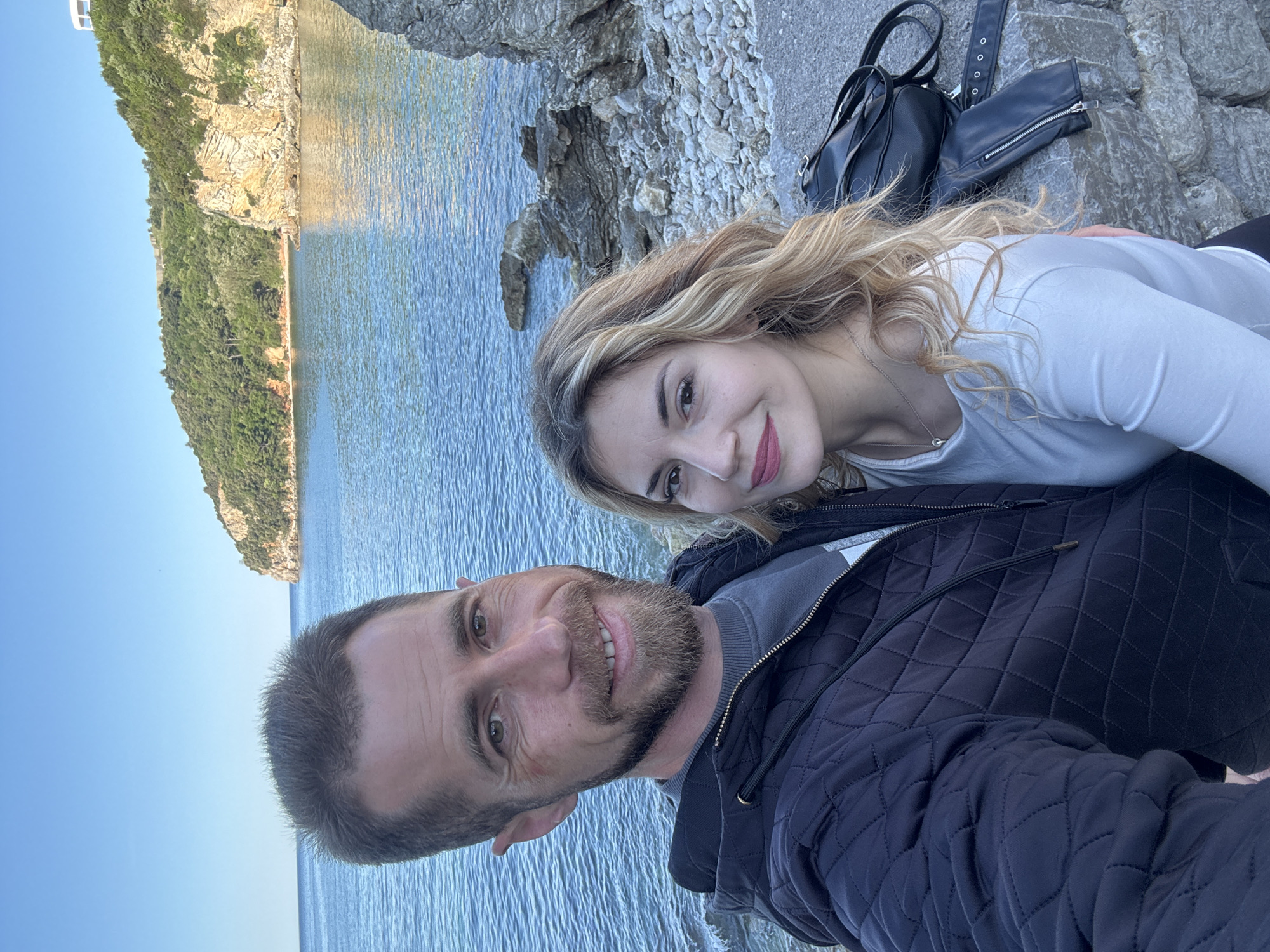 A couple poses for a selfie on rocks near the sea. The man is smiling and wearing a dark, quilted jacket. The woman has long blonde hair and a white shirt. A black backpack and a piece of black clothing are on the rocks behind them. The sea and a cliff covered with trees are in the background. The location is most likely in or near the city of Budva, Montenegro.