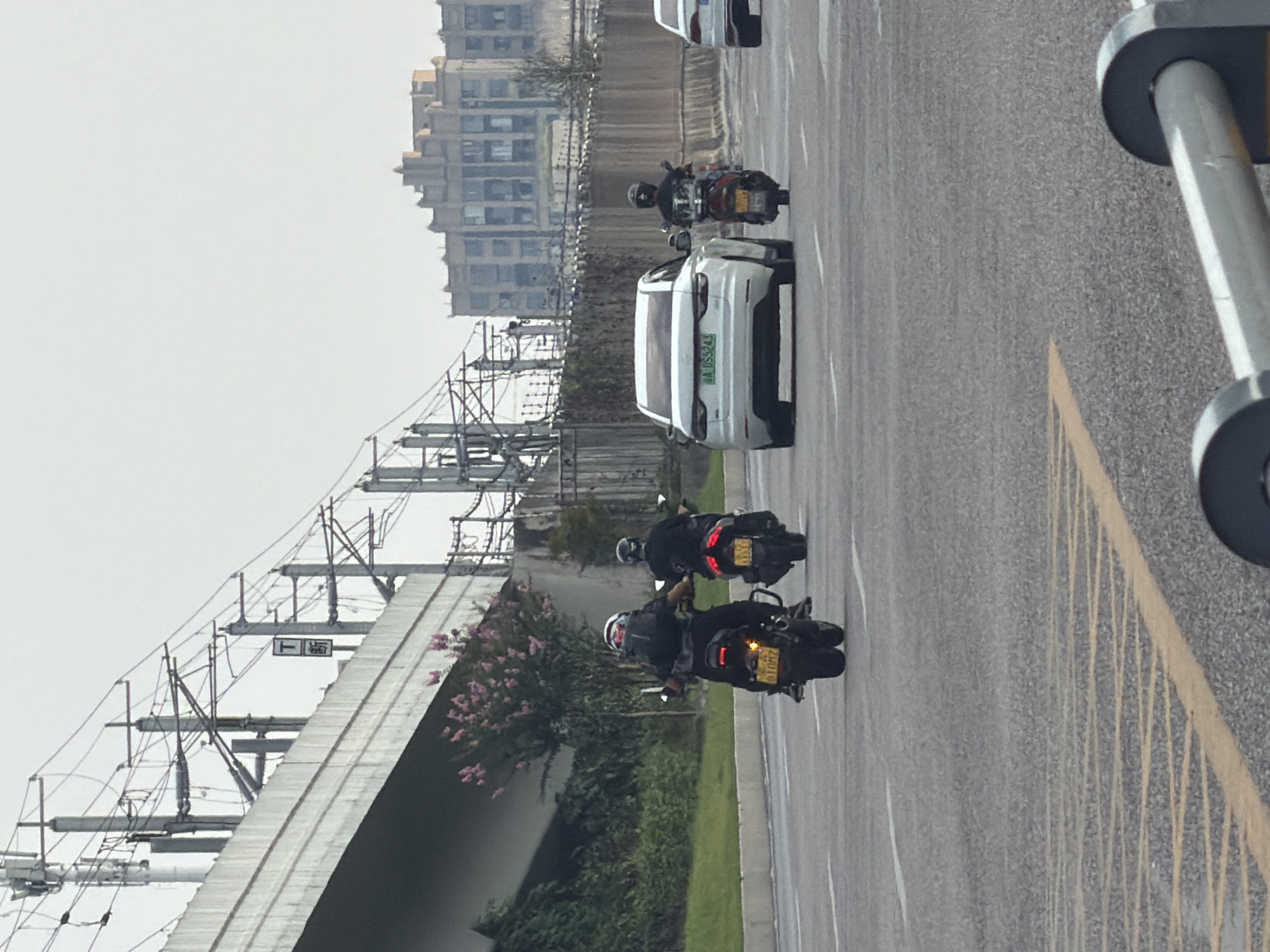 The image shows a street scene in Jiangjin, China, with several vehicles on a paved road under an overpass. In the foreground, two motorcycles are riding away from the camera. The rider of the first motorcycle, on the left, is wearing a backpack and a helmet. The second motorcycle, slightly behind and to the right, also has a rider with a helmet. Further down the road, a white car is moving away, and to its right, another motorcycle with a rider is visible. In the background, there is an elevated structure with electrical wires and poles, and beyond that, a multi-story building with a distinctive facade. The environment appears to be an urban setting with traffic and infrastructure.