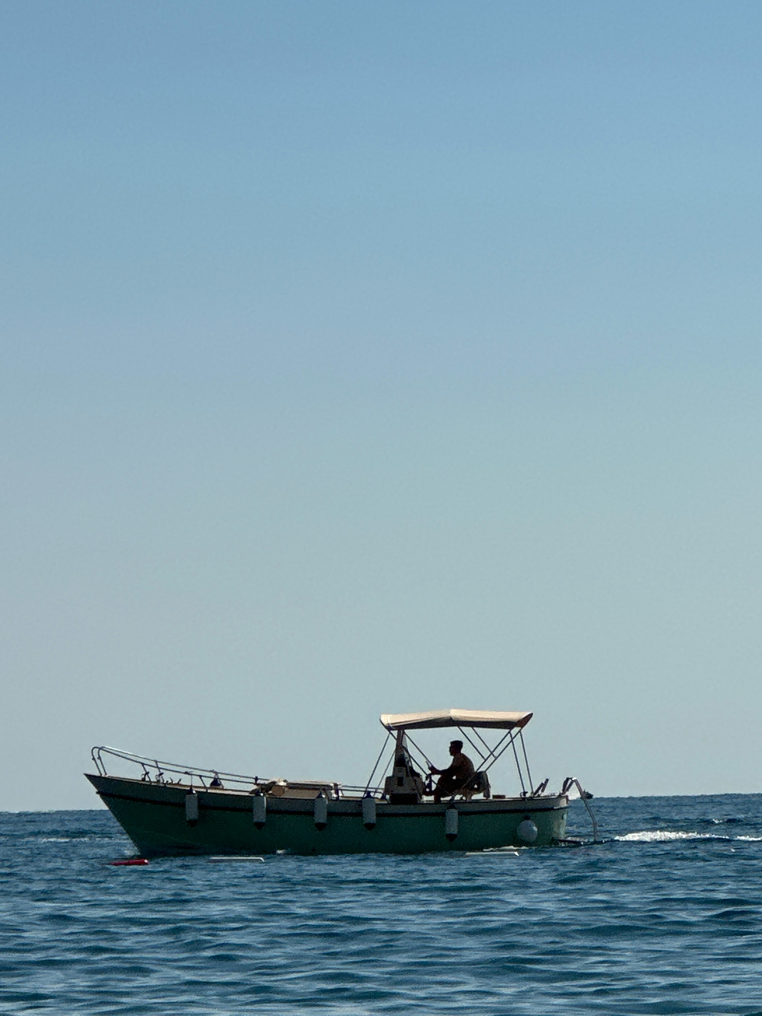 A small, green boat with a white canopy is moving through blue, choppy water under a clear sky. A lone individual, silhouetted, is seated at the helm of the boat, appearing to steer. The boat has a metal railing along its edge and several fenders attached. The scene suggests a calm day at sea, possibly near the coast of Boreti, Montenegro, given the clear blue sky and the presence of a boat on the water.