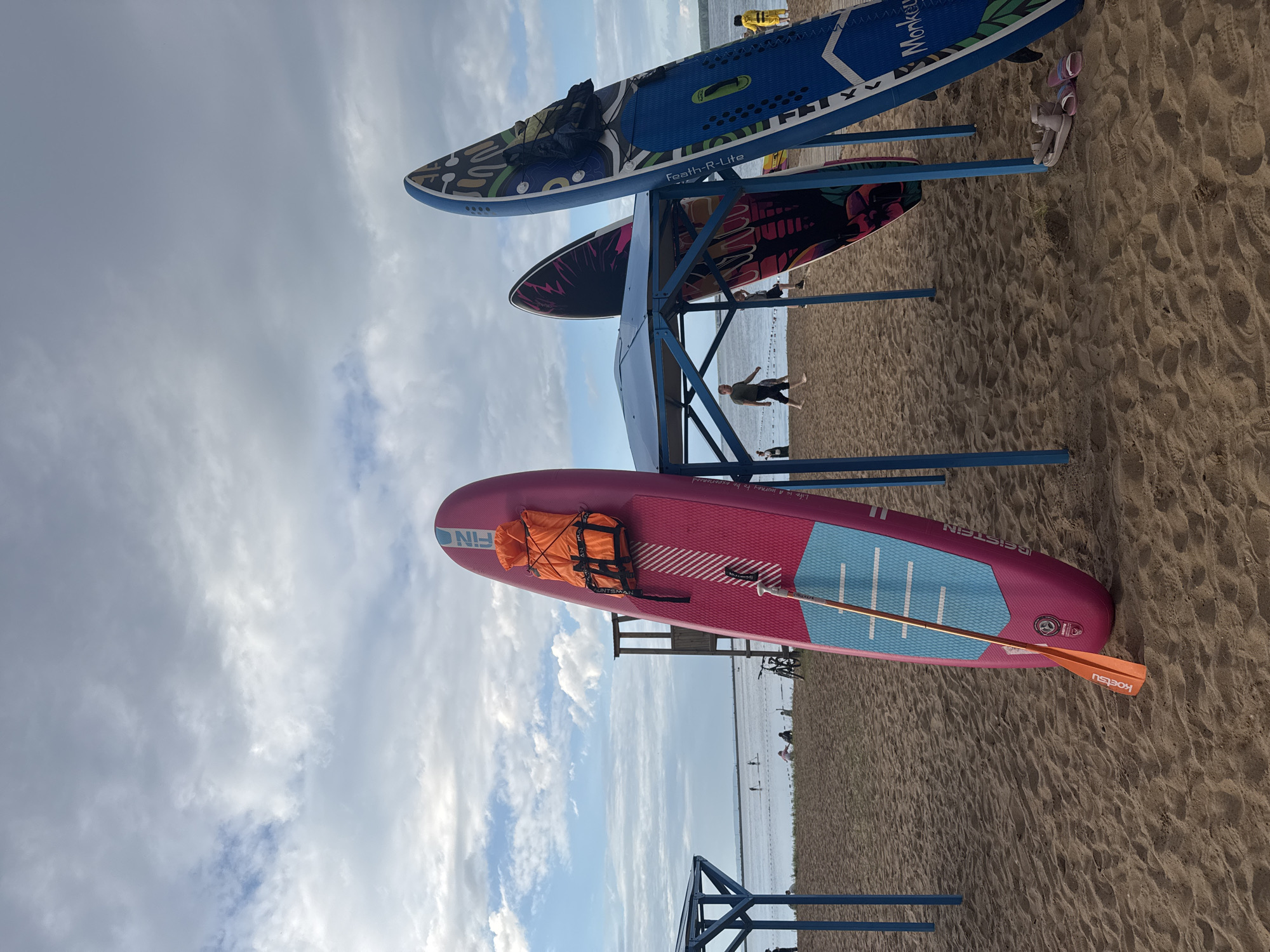 This media file shows a collection of paddleboards neatly stored on a sandy beach. The setting is a beach with calm water in the background, suggesting a lake or a very still sea. The sky is partly cloudy, with a mix of white and gray clouds, indicating it's likely daytime, perhaps late afternoon given the soft light.
There are at least three paddleboards visible on a blue metal rack. The top paddleboard is blue with a colorful, abstract design and text that appears to say "Feath-R-Lite." Below it is a paddleboard with a black and pink floral design. The paddleboard closest to the viewer is bright pink and features a blue traction pad, an orange life vest secured with straps, and a paddle with an orange blade resting on it. Text visible on the pink paddleboard includes "FIN" and "BEISTEIN," and on the paddle it says "KOETSU."
In the background, several people are visible near the water's edge, some standing and others possibly walking or swimming. There are also bicycles parked on the sand. The overall mood is relaxed and suggests a recreational setting where people are preparing for or have just finished water activities. The sand appears to be damp in some areas, and the presence of footprints indicates recent activity. The scene is set under a moderately cloudy sky, with the sun filtering through, creating a soft illumination.