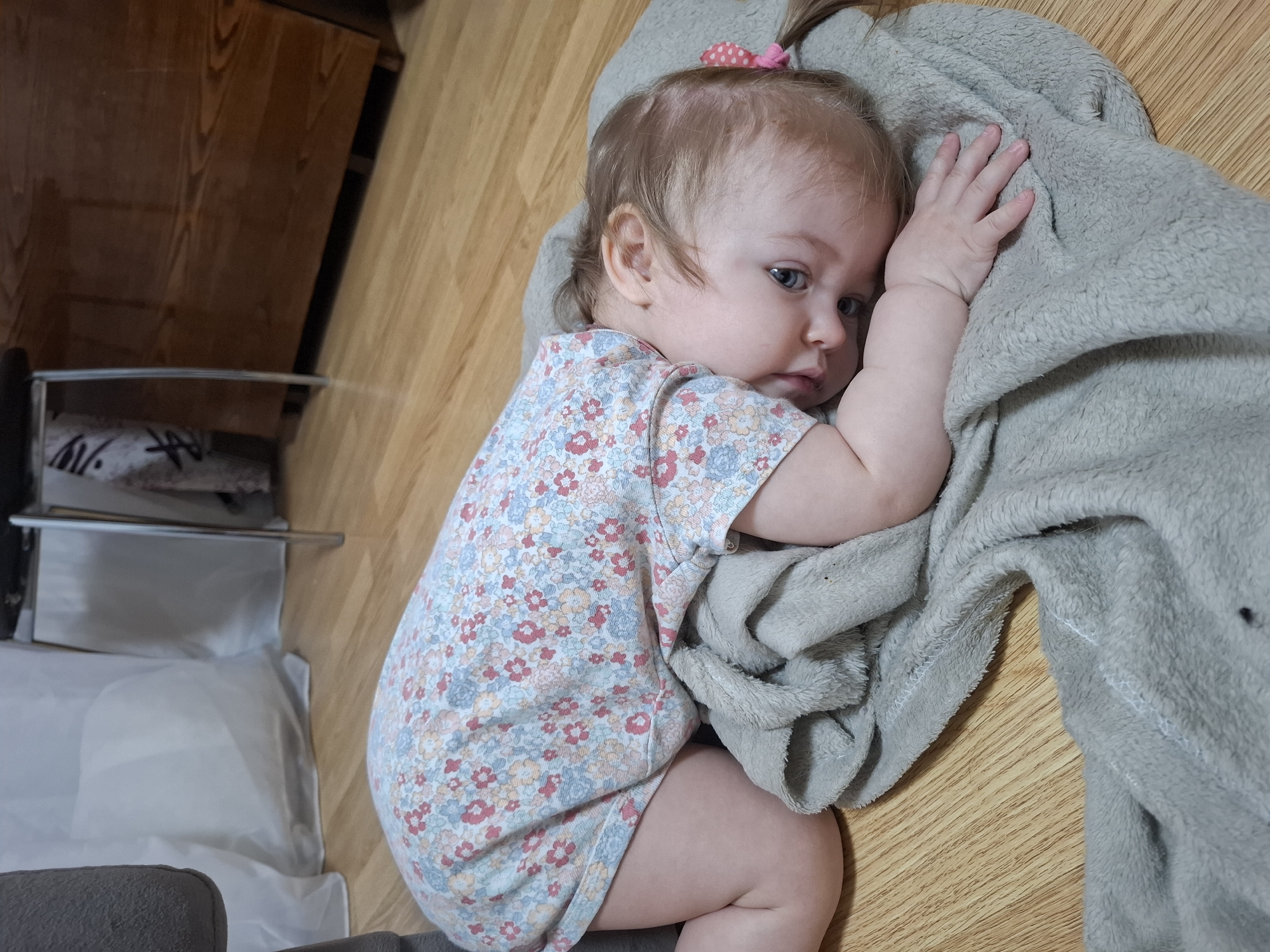 A baby wearing a floral onesie is lying on a wooden floor, partly covered by a light grey blanket. The baby has fair skin, blue eyes, and light brown hair with a small pink hair tie. The baby is lying on its side, looking to the right, with its left arm extended and hand resting on the blanket. To the left of the baby, there's a piece of furniture with a shelf visible. The location is likely a home. There is no confirmed information of this image being taken in Chaikovsky, Russia.