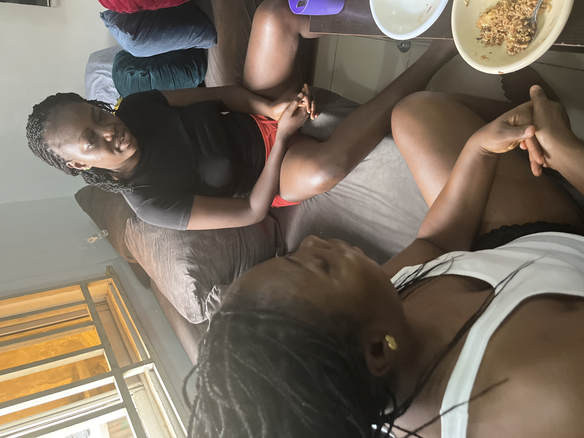 The image depicts an indoor scene, possibly a living room, in Abuja, Nigeria. Two young Black women are seated on a couch. The woman on the right is wearing a black t-shirt and red shorts, with her hands clasped in her lap. The woman on the left is wearing a white tank top and black shorts, and her arms are resting in her lap. There is a table in front of them with two bowls, one containing rice and the other appearing empty. The background shows a window with bars and a light-colored wall. The overall impression is one of casual conversation or relaxation.