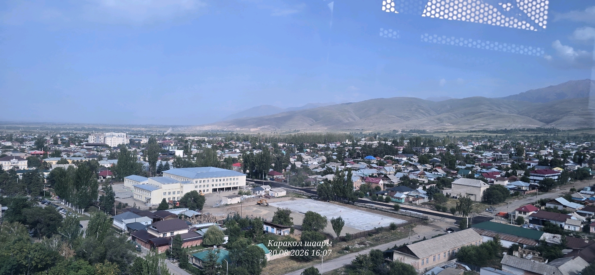 The image is an aerial view of a city with mountains in the background, likely taken from a high vantage point. The city is densely populated with residential buildings of various sizes and architectural styles, characterized by pitched roofs in different colors, including red, blue, and grey. There are also larger, multi-story institutional buildings with light-colored facades. Green trees are interspersed throughout the urban landscape, and some areas show signs of ongoing construction or development, with exposed ground and construction equipment visible. The sky is clear and light blue. A watermark in the lower right corner reads "Каракол шаары 30.08.2026 16:07", indicating the location as Karakol City, Kyrgyzstan, and the date and time as August 30, 2026, at 16:07. The reflection of a patterned ceiling or structure is visible in the top portion of the image, suggesting the photo was taken through a window.