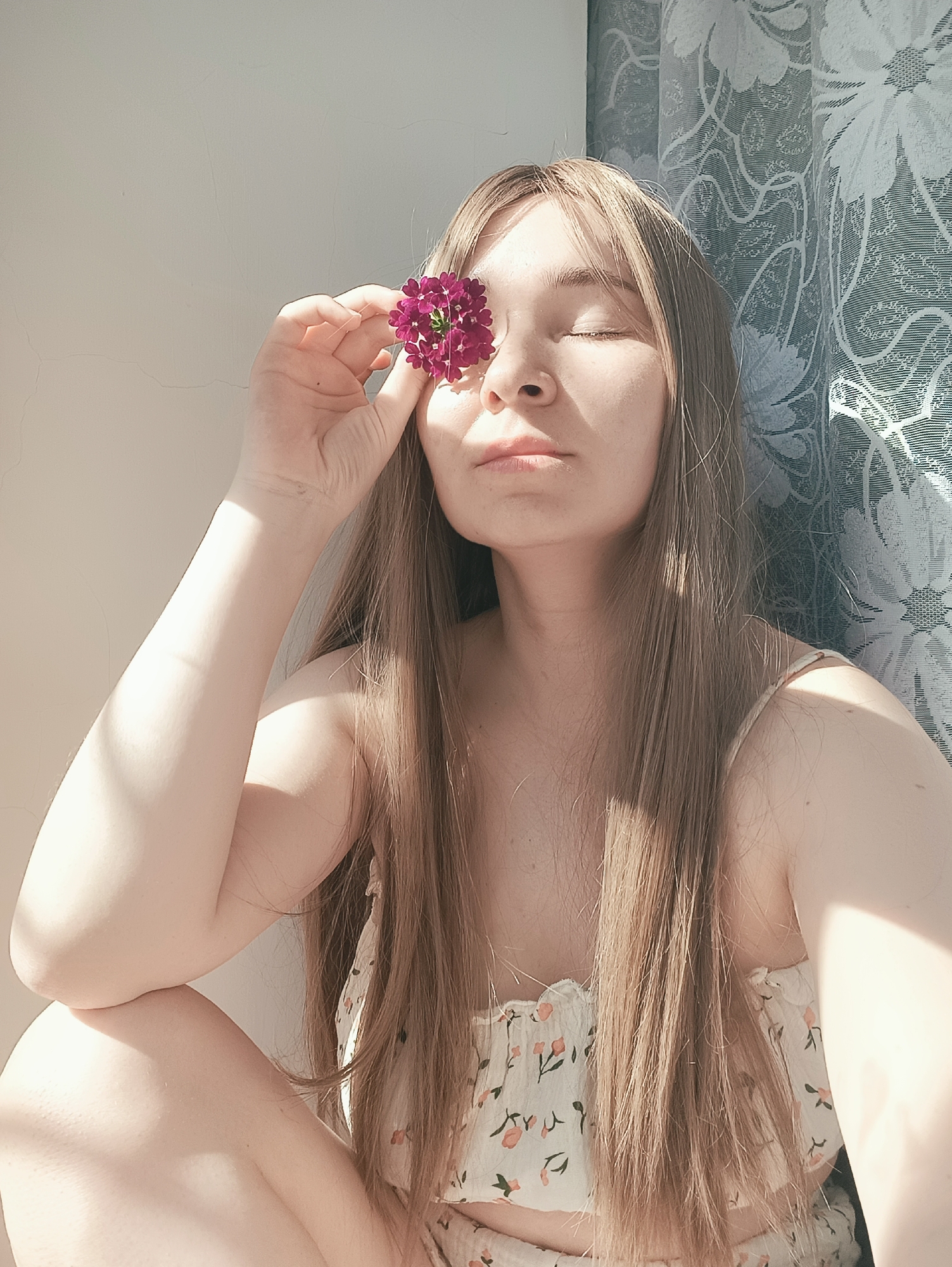 A fair-skinned woman with long, straight, light brown hair sits indoors near a window. She has her eyes closed and is holding a purple flower in front of one eye. She is wearing a floral-patterned, light-colored top with spaghetti straps. The background includes a plain white wall and a window with a patterned lace curtain. The scene is illuminated by sunlight. The image does not depict any information specific to the city of Izhevsk, Russia.