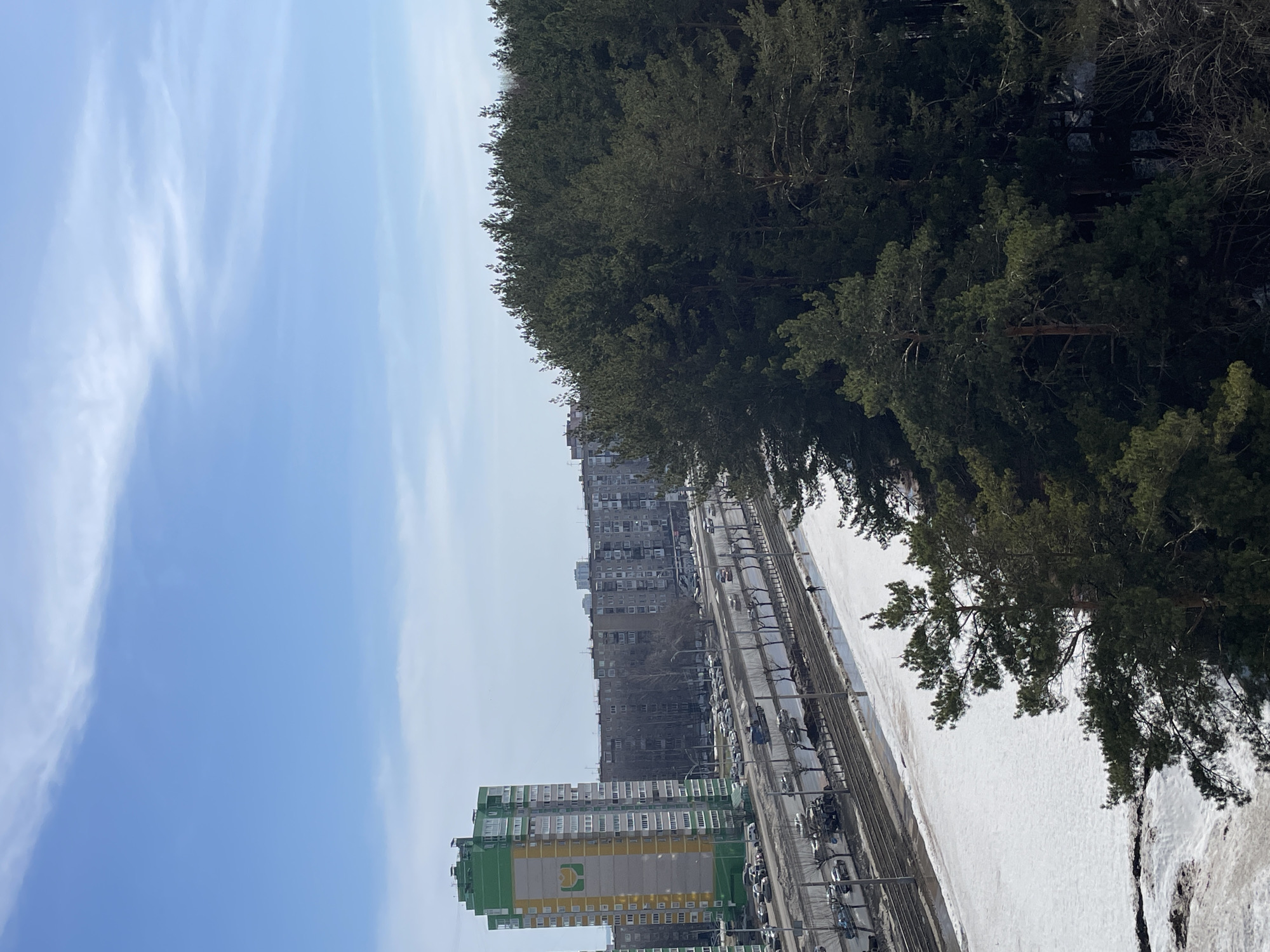 The image shows an urban scene in Izhevsk, Russia, with a view of residential buildings, a road with cars, a patch of snow, trees, and a blue sky with clouds.
In the foreground, there is a tall, green and yellow apartment building with a logo visible, likely a grocery store or other commercial establishment. A road runs alongside the building, filled with parked cars. A second row of buildings stands behind the first, appearing older and gray.
A large patch of snow extends from the road toward the right side of the image. Trees are present on the right, obscuring some of the snow and adding a natural element to the scene.
The sky is blue with wispy clouds.