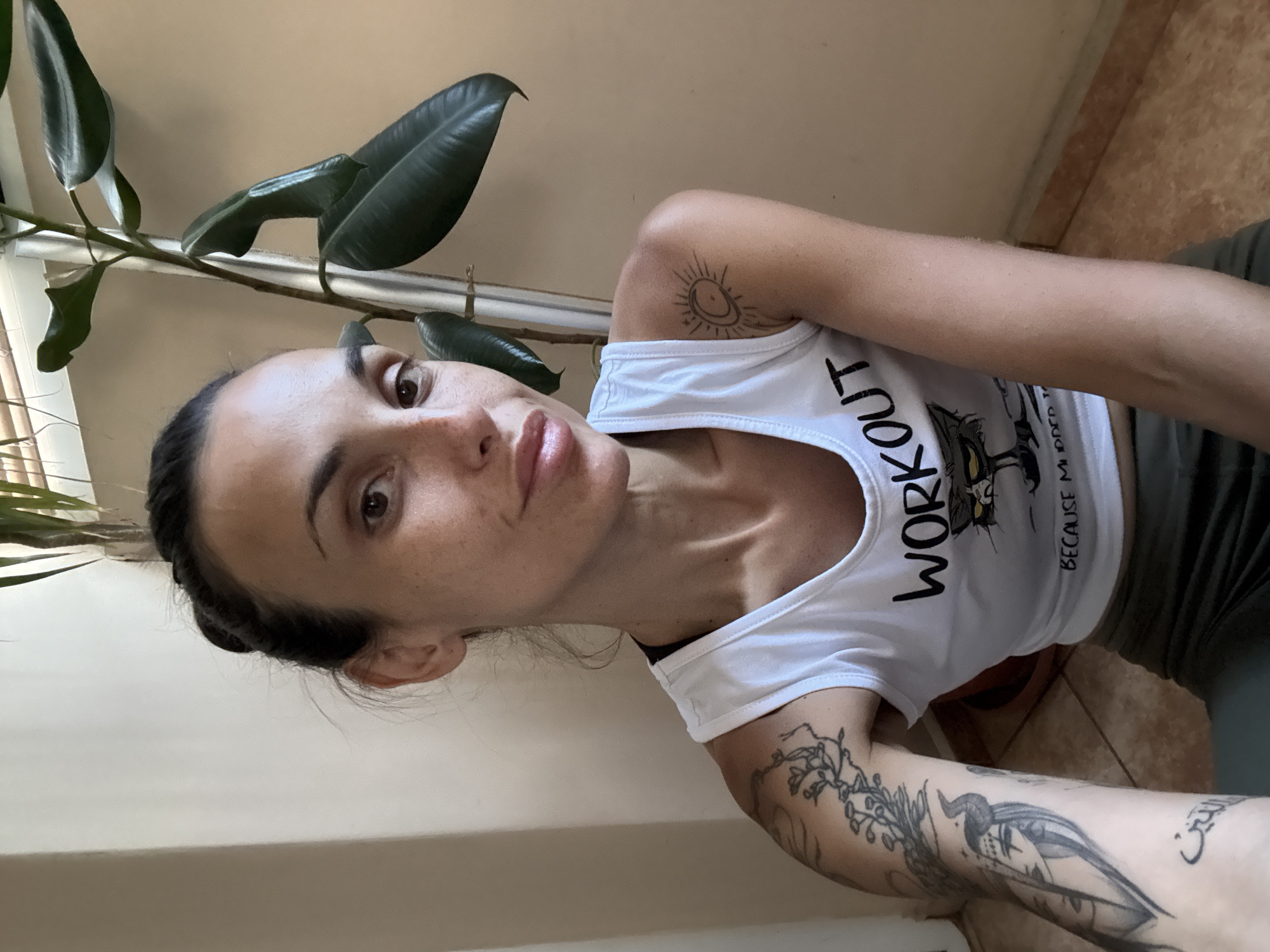 A woman with dark hair pulled back in a bun, wearing a white tank top with a "WORKOUT BECAUSE MURDER IS WRONG" graphic and olive green leggings, poses for a selfie indoors. She has multiple tattoos on her arms, including a detailed depiction of a horned figure and floral designs. A large potted plant with broad green leaves is visible to her right. The background consists of a neutral-colored wall and tiled flooring. The image was taken in Pleven, Bulgaria.